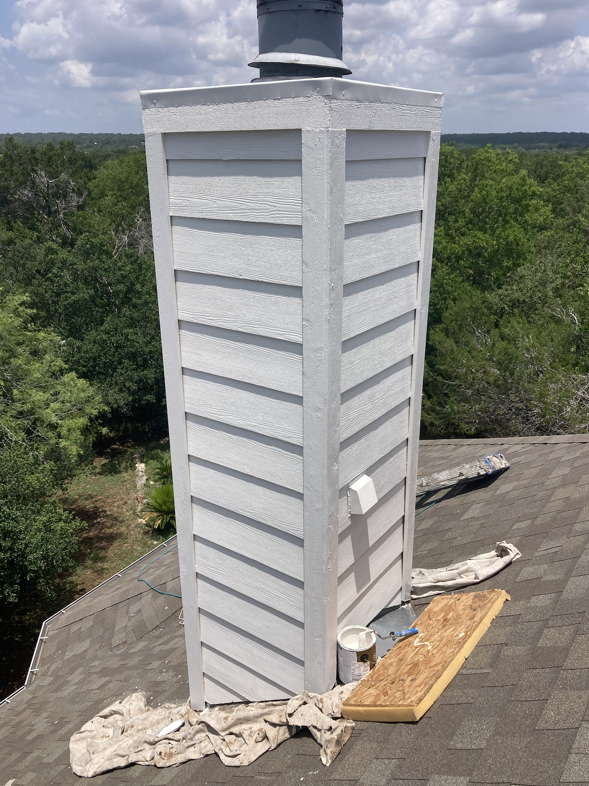 A new white-painted chimney on a residential rooftop with some construction supplies and cloth around its base, surrounded by trees under a partly cloudy sky.