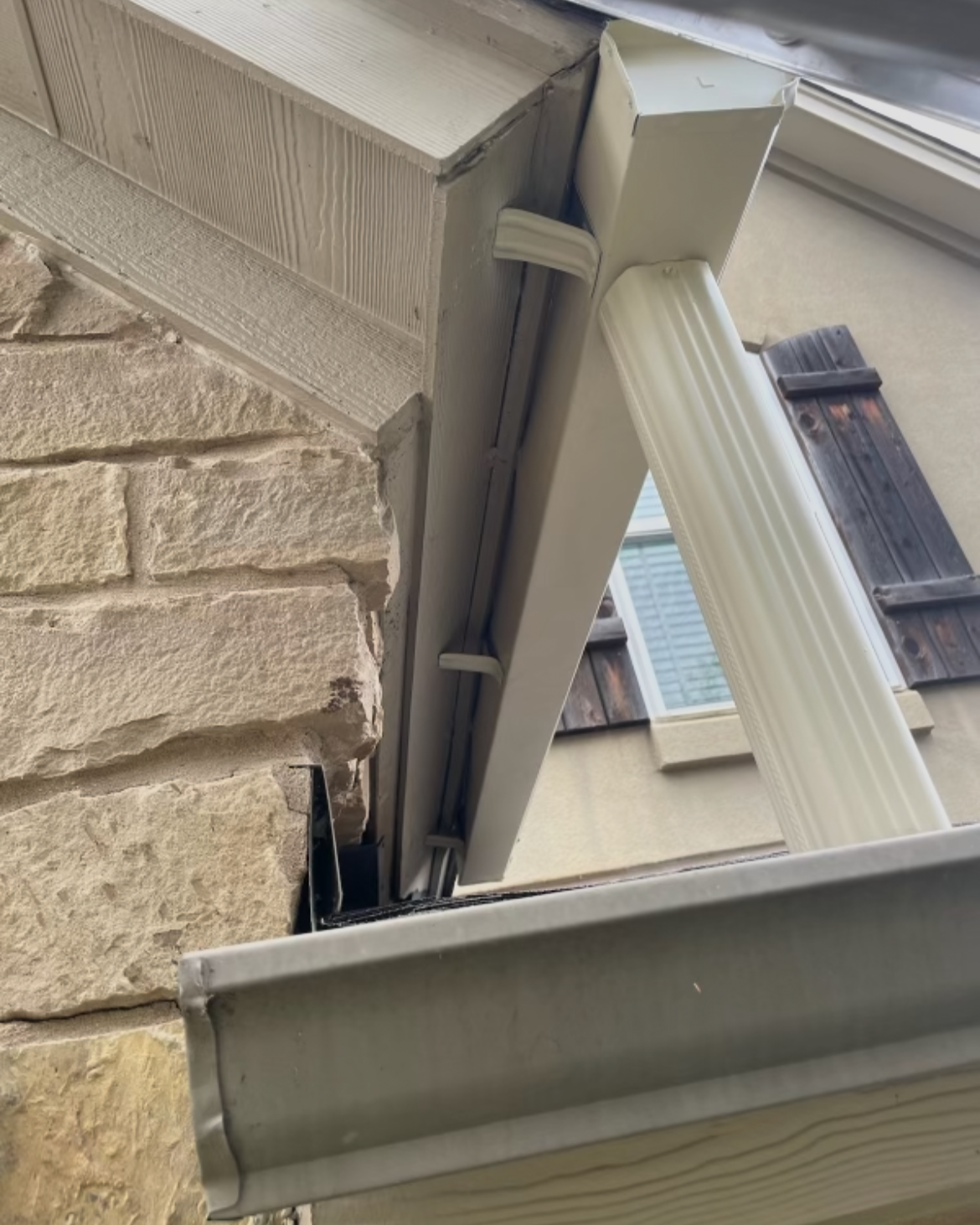 Close-up of a house exterior corner showing stone and siding, with a window frame and a white decorative column, and a part of the house roof with shingles.