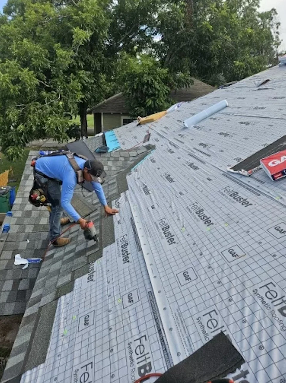 A worker on a roof installing new shingles, with roofing underlayment visible and trees in the background.