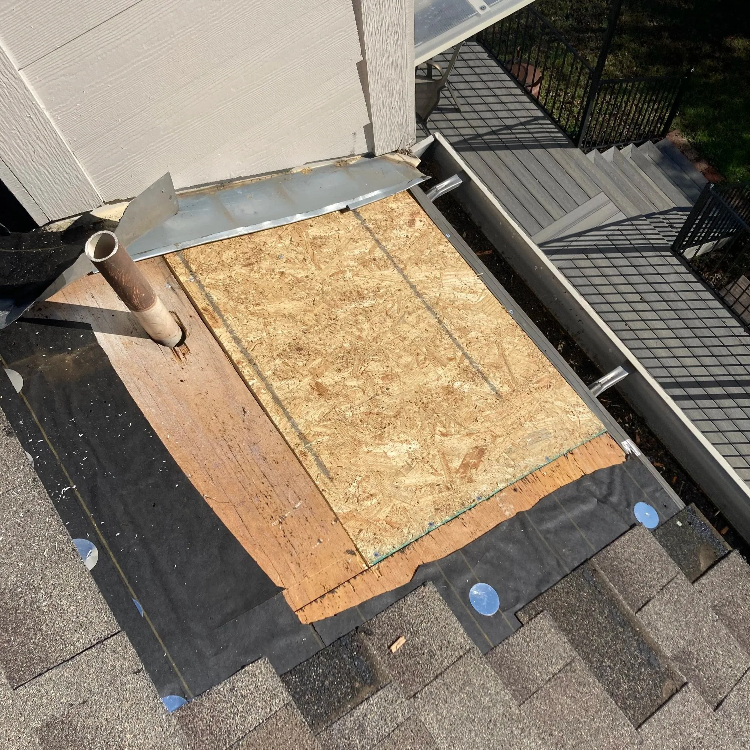 A section of a house exterior under construction showing roofing with black felt paper, a plywood section, and metal flashing along the edge of the roof. Roof drainage pipes and rooftop stairs are visible.