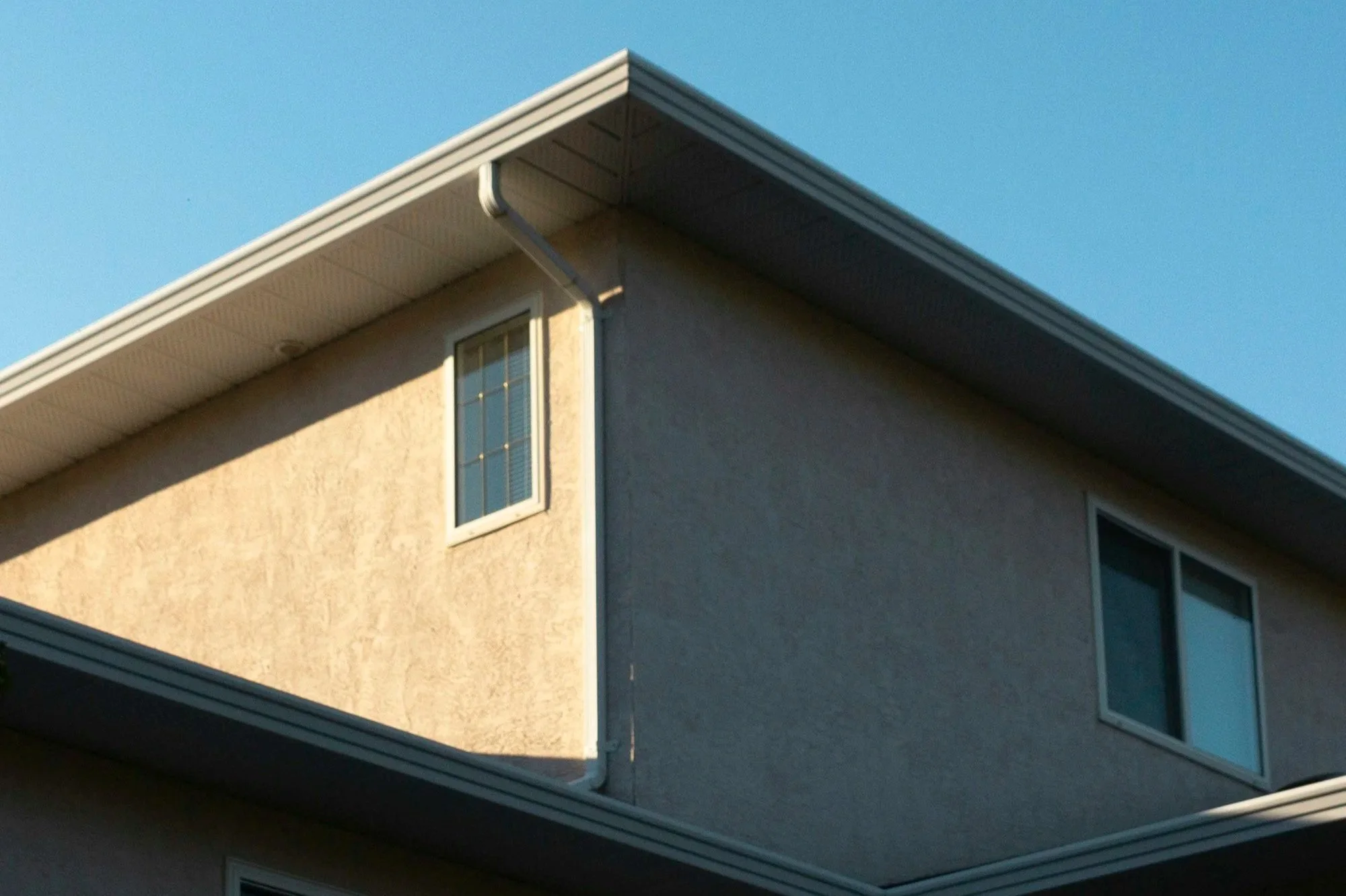 Close-up view of the corner of a house showing beige and gray stucco walls, a window with multiple small panes, and a roof with white gutters and dark shingles under a clear blue sky.