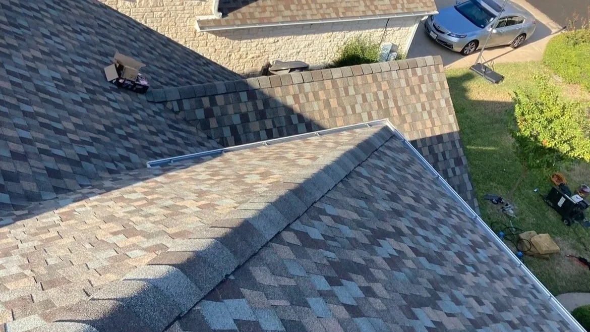 View from a roof showing shingles, adjacent rooftops, a driveway with a parked silver car, a grassy yard, and gardening tools.