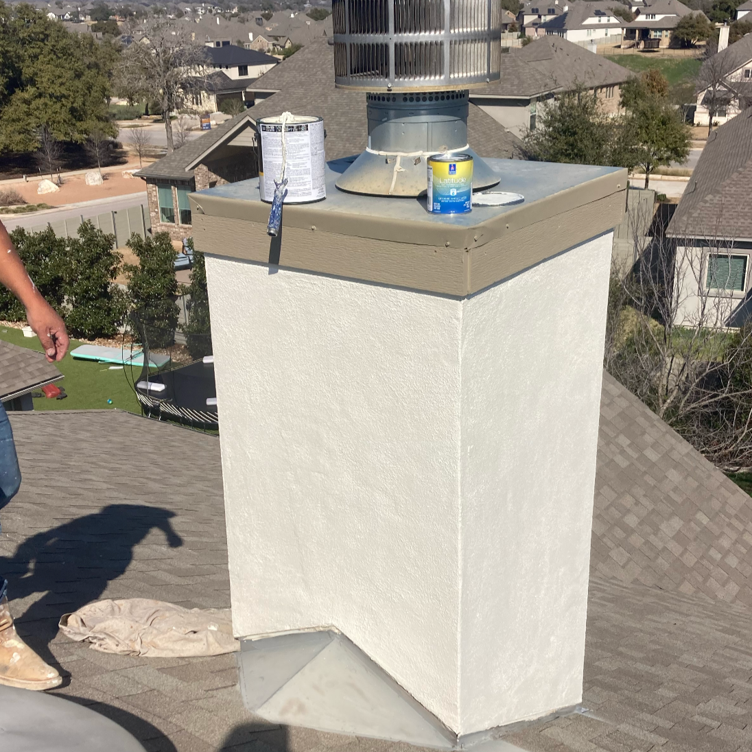 Close-up of a chimney on a residential roof, with cans of rust-inhibiting paint and a small paintbrush on top, and a black gutter running along the roof edge.