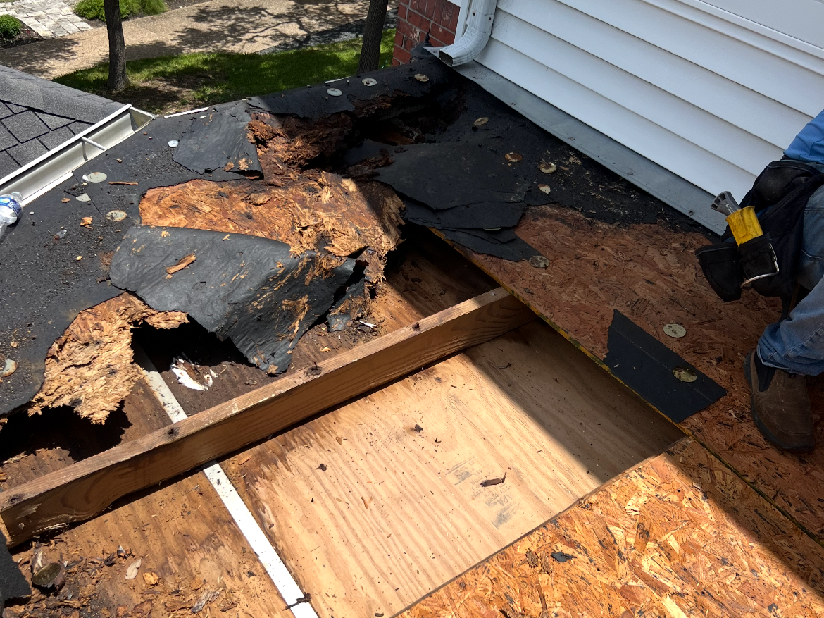 Roof damage showing a section of asphalt shingles and underlayment removed, revealing wood decking and support beams underneath, with roofing materials and tools nearby.