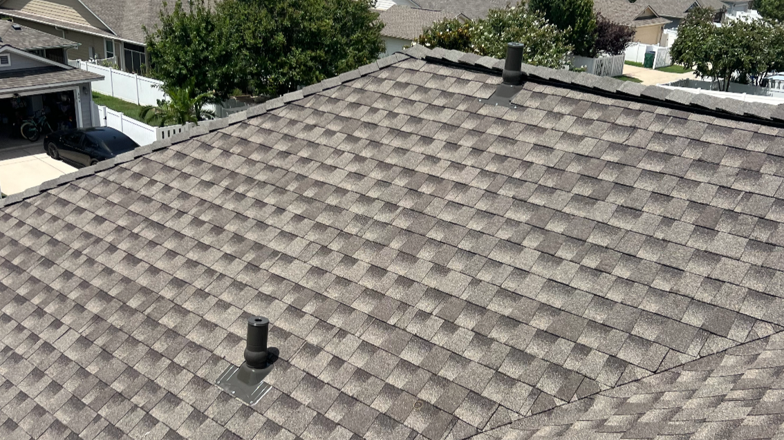 View of a residential roof with gray asphalt shingles, featuring two black vents, surrounded by trees and neighboring houses.