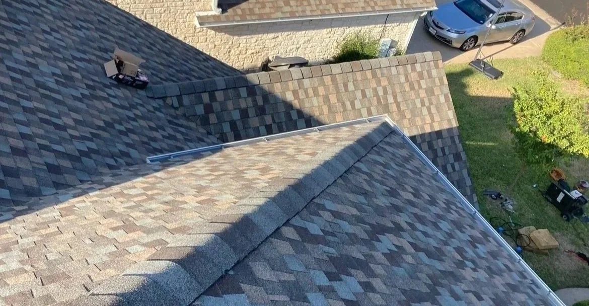 Aerial view of a house roof with multiple sections of asphalt shingles, a nearby driveway with a silver car, and a yard with trees and equipment.