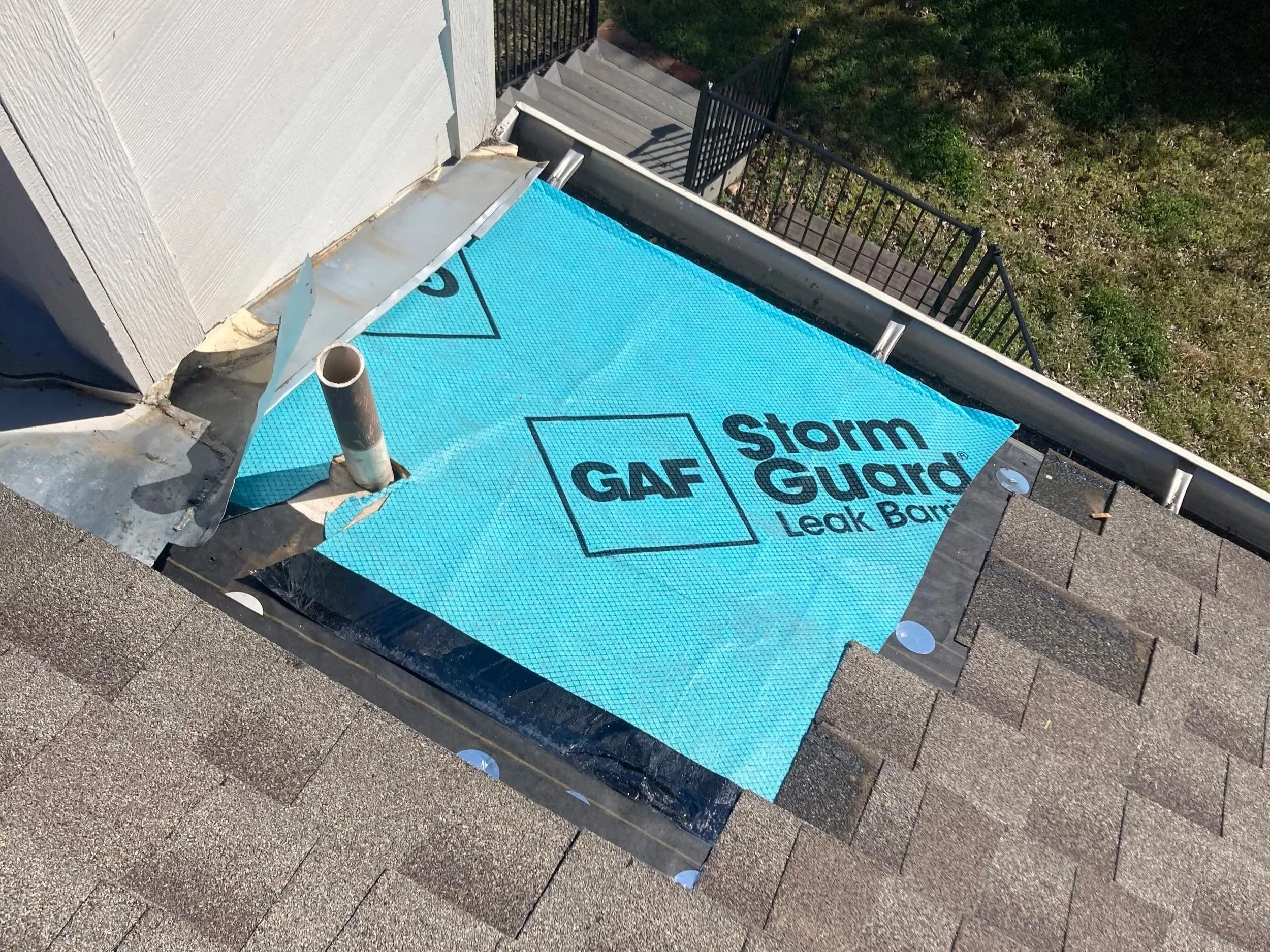 View of a roof with a blue StormGuard Leak Barrier underlayment partially installed near the edge, adjacent to a gutter and a black metal railing on a backyard with green grass.