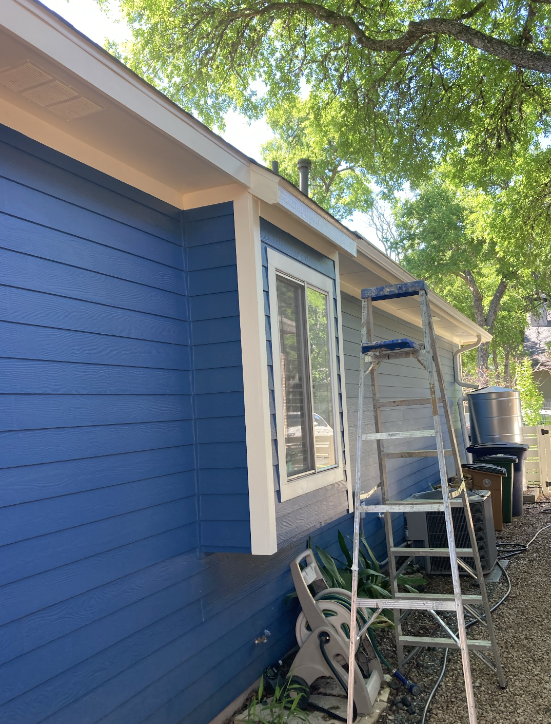 Side of a house with blue siding, a window, a metal ladder, and outdoor equipment, with trees in the background.