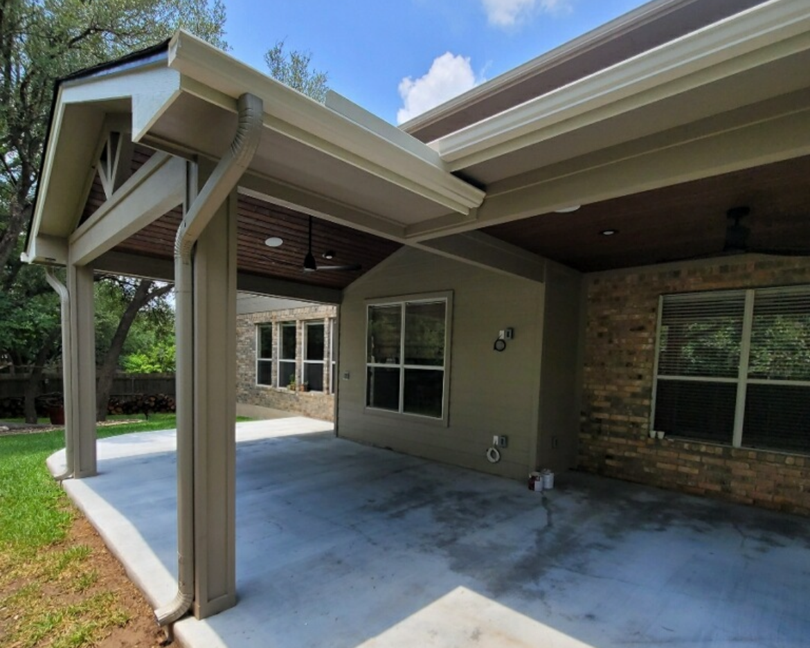 Backyard patio with a concrete slab, covered by a roof extension, with a brick and siding house wall, windows, and trees in the background.