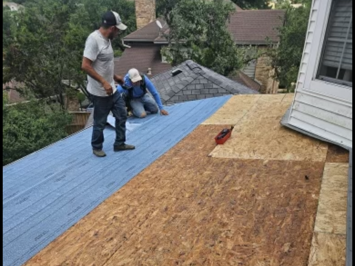 Two men working on a roof during a roofing project. One man is standing, the other is kneeling, both are installing or inspecting roofing materials. The roof is partially covered with blue underlayment, with areas of exposed plywood, in a residential neighborhood with trees and houses in the background.