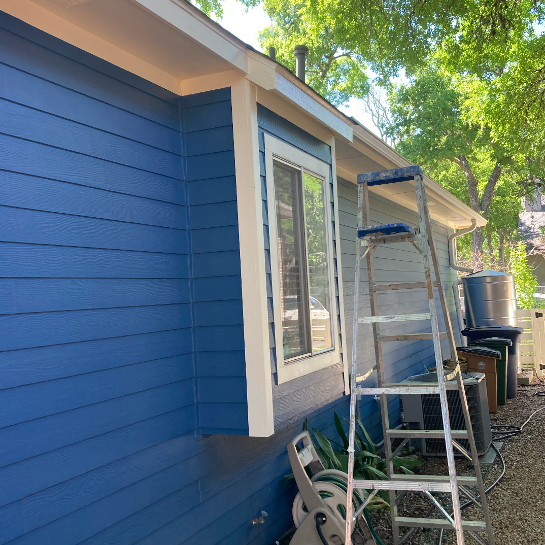 Blue house exterior wall with a window, ladder, and gardening tools visible, surrounded by trees and outdoor equipment.