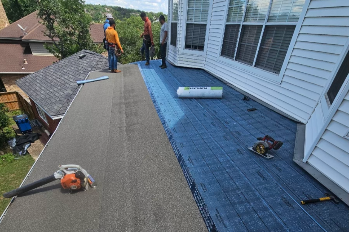 Construction workers installing a new roof on a house, with some workers standing on the roof and equipment laid out, including a circular saw and roll of roofing underlayment.
