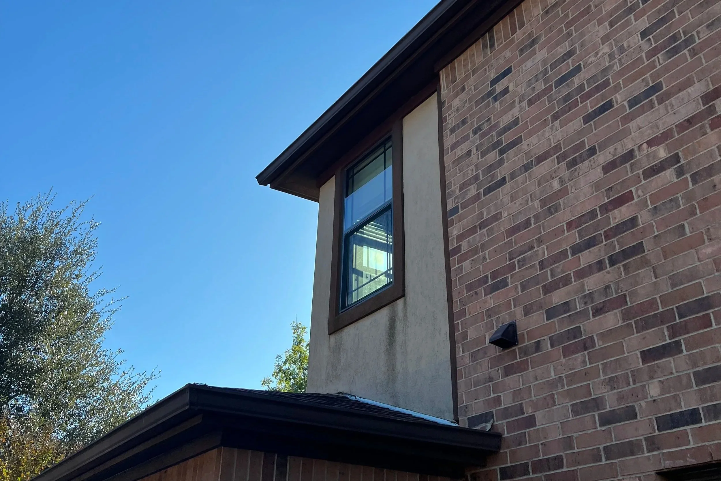 Close-up view of a brick house corner with a window and a dark roof, under a clear blue sky with some trees visible in the background.
