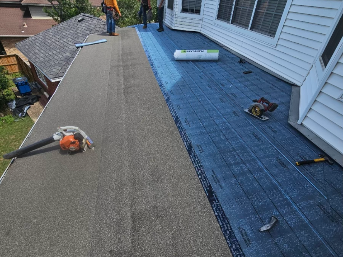 Roof undergoing construction with new roofing materials being installed. Tools and materials are visible on the roof, with workers working near the edge. The roof has a section with weatherproof underlayment and another section with finished shingles.