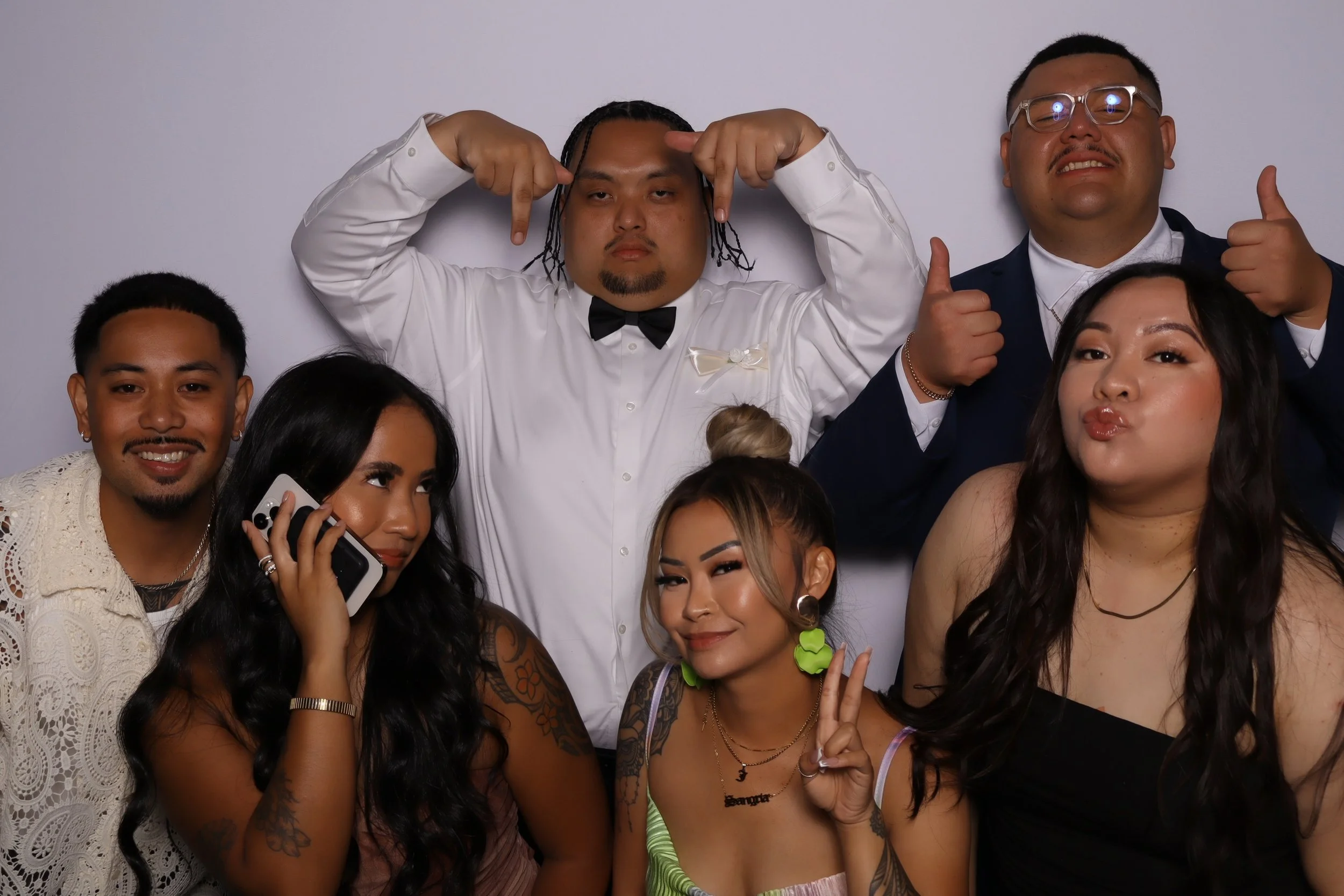 Group of six diverse young adults posing together at a party, with some making gestures, smiling, and one woman talking on her phone. They are dressed in stylish casual and semi-formal outfits, with a plain gray background.