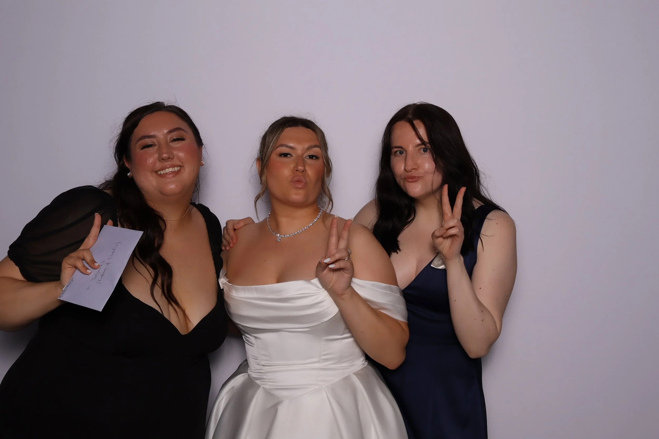 Three young women at a formal event, standing close together and making peace signs with their hands, with a plain white wall in the background.