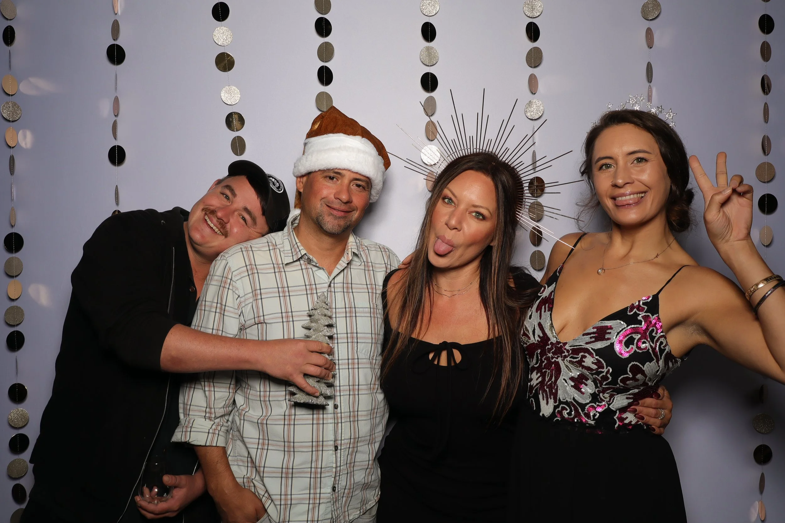 Group of four friends celebrating Christmas, wearing festive accessories and posing playfully in front of a decorated backdrop.