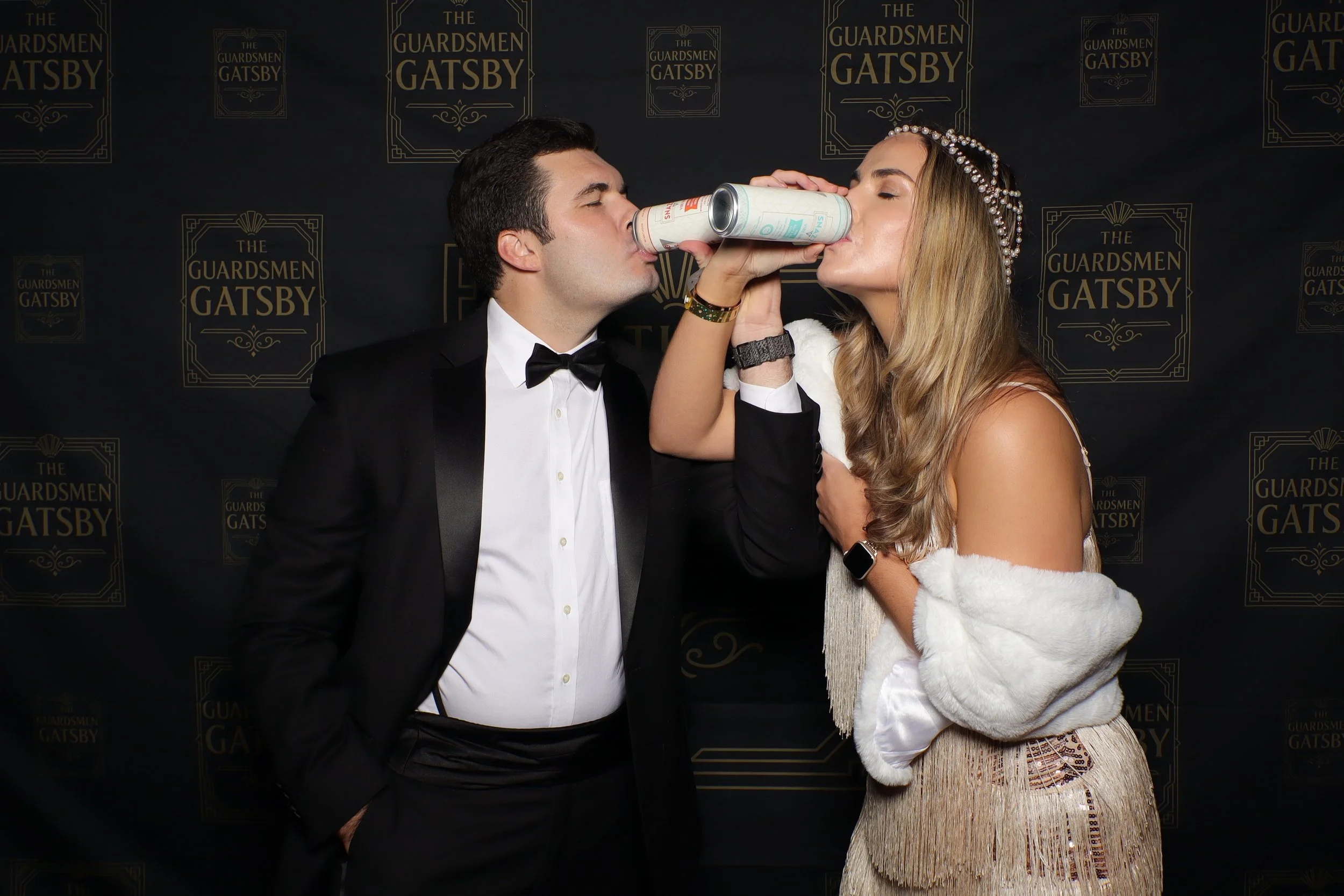 A man and woman in formal attire sharing a toast at a Gatsby-themed event, with the woman wearing a pearl headband and the man in a tuxedo.