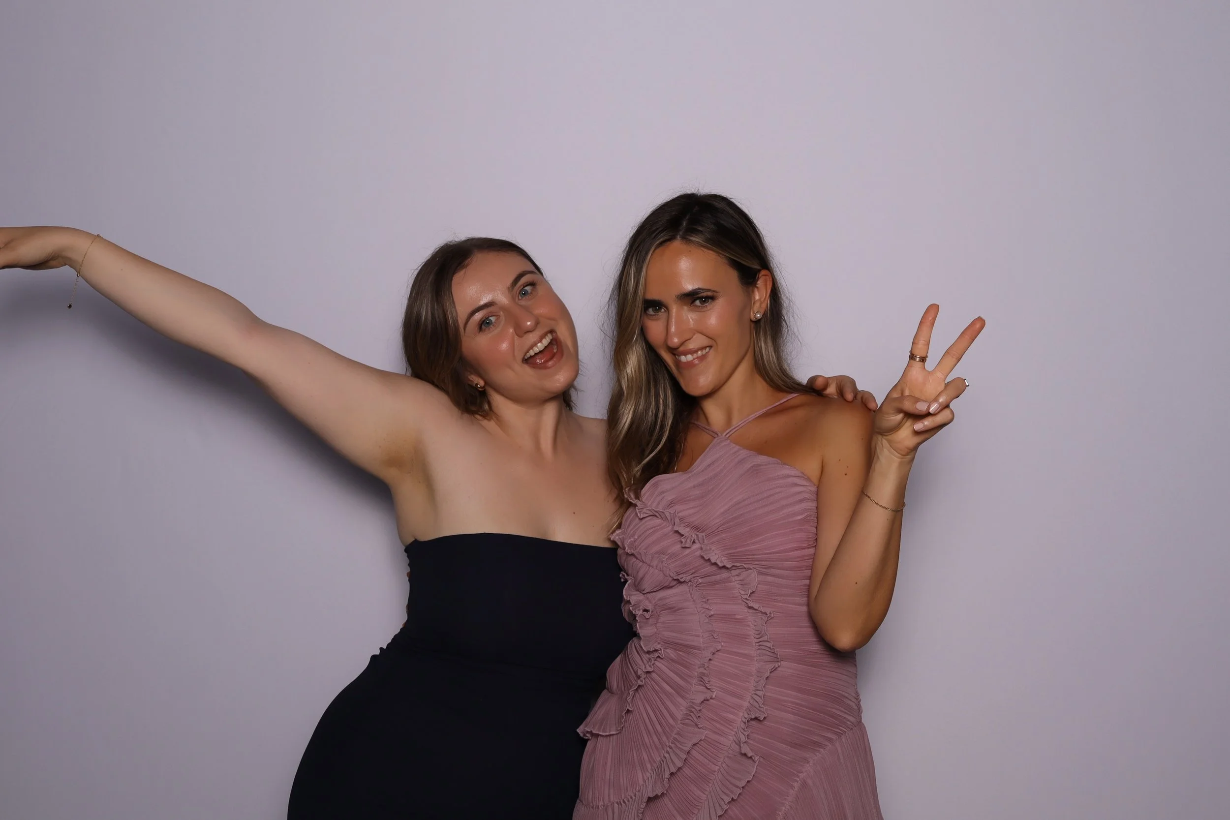 Two women smiling and posing for a photo against a plain wall, one making a peace sign and the other extending an arm.