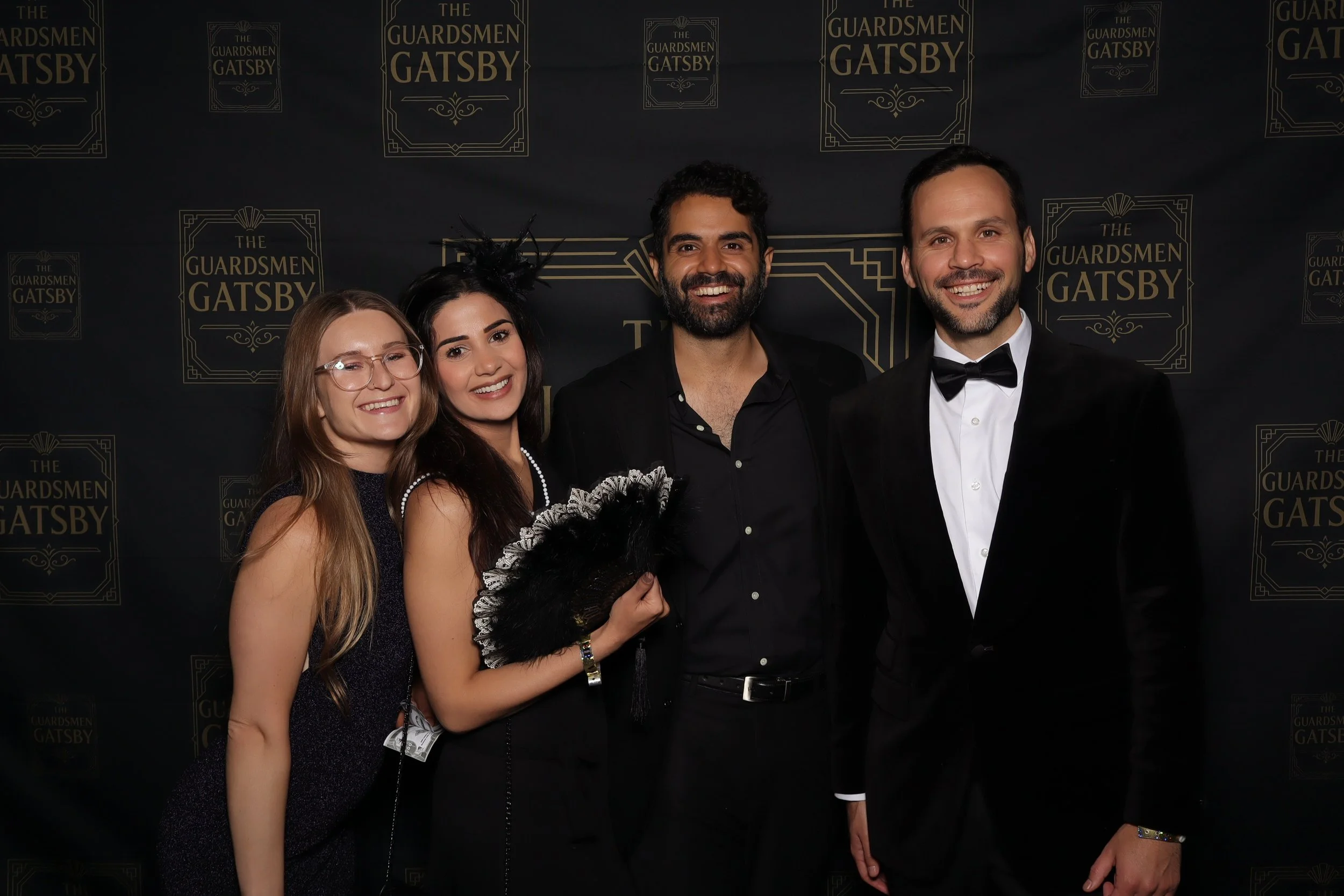 Group of four people dressed in formal attire, standing in front of a black backdrop with the text 'The Guardsmen Gatsby' repeatedly printed, smiling at the camera at a themed event or party.