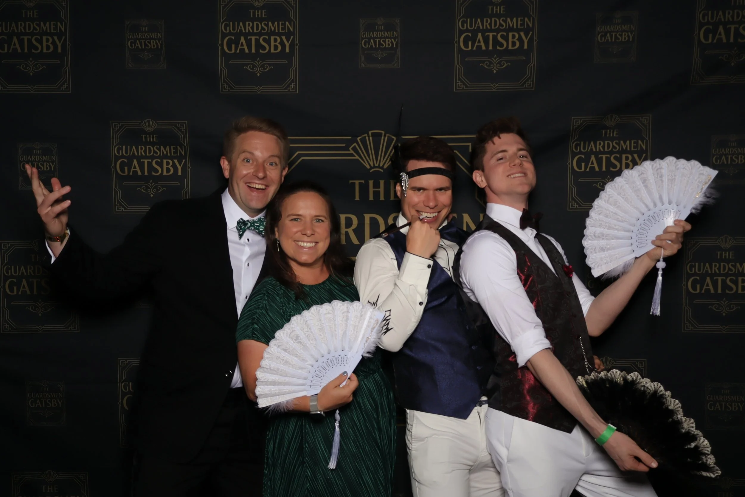 Five people at a Gatsby-themed event posing in front of a black backdrop with gold text that reads 'The Guardsmen Gatsby', smiling, some holding decorative fans.