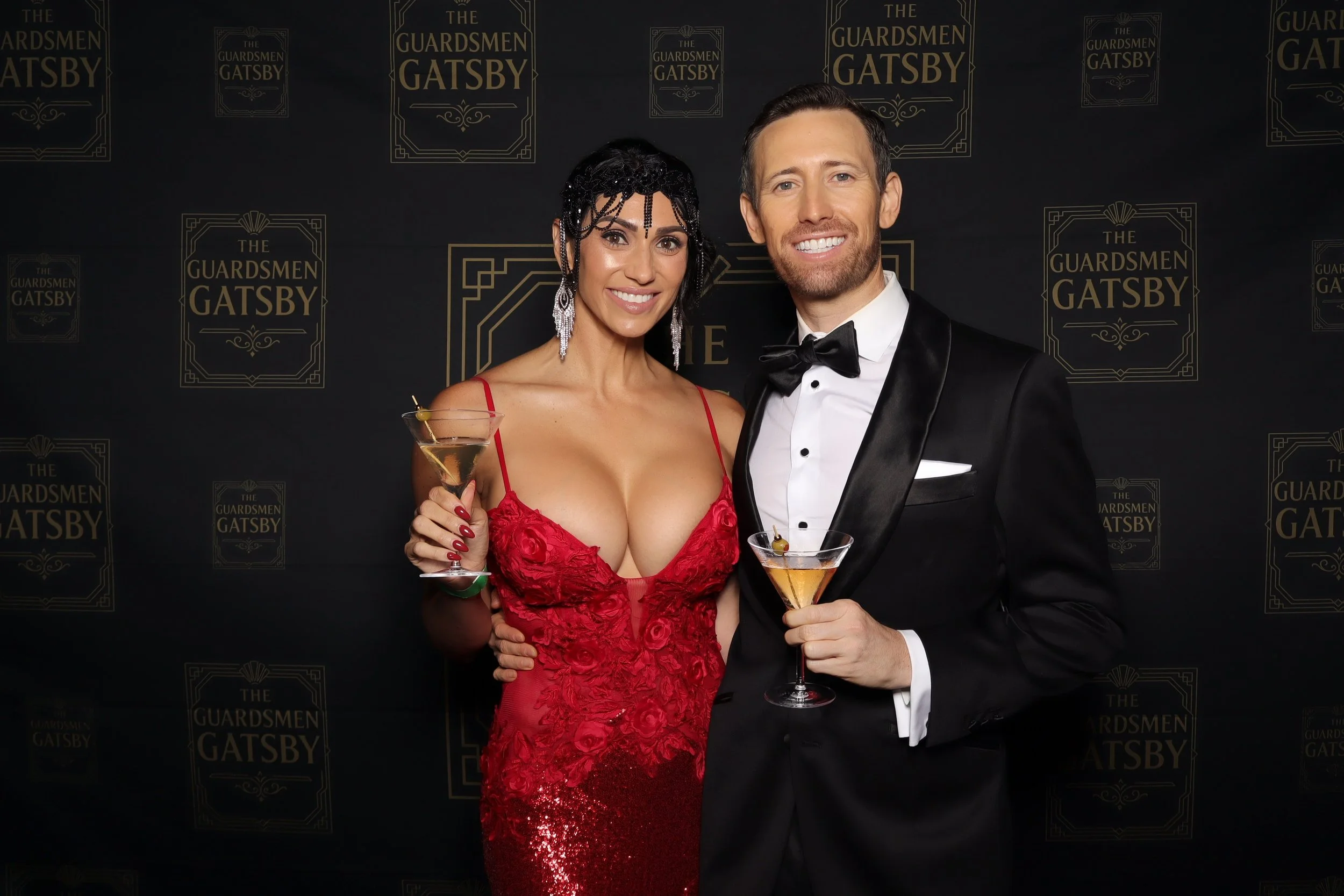 A woman in a red dress and a man in a black tuxedo with a bow tie are smiling and holding cocktails at an event celebrating 'The Guardsmen Gatsby,' with a black backdrop featuring this name repeatedly in gold.