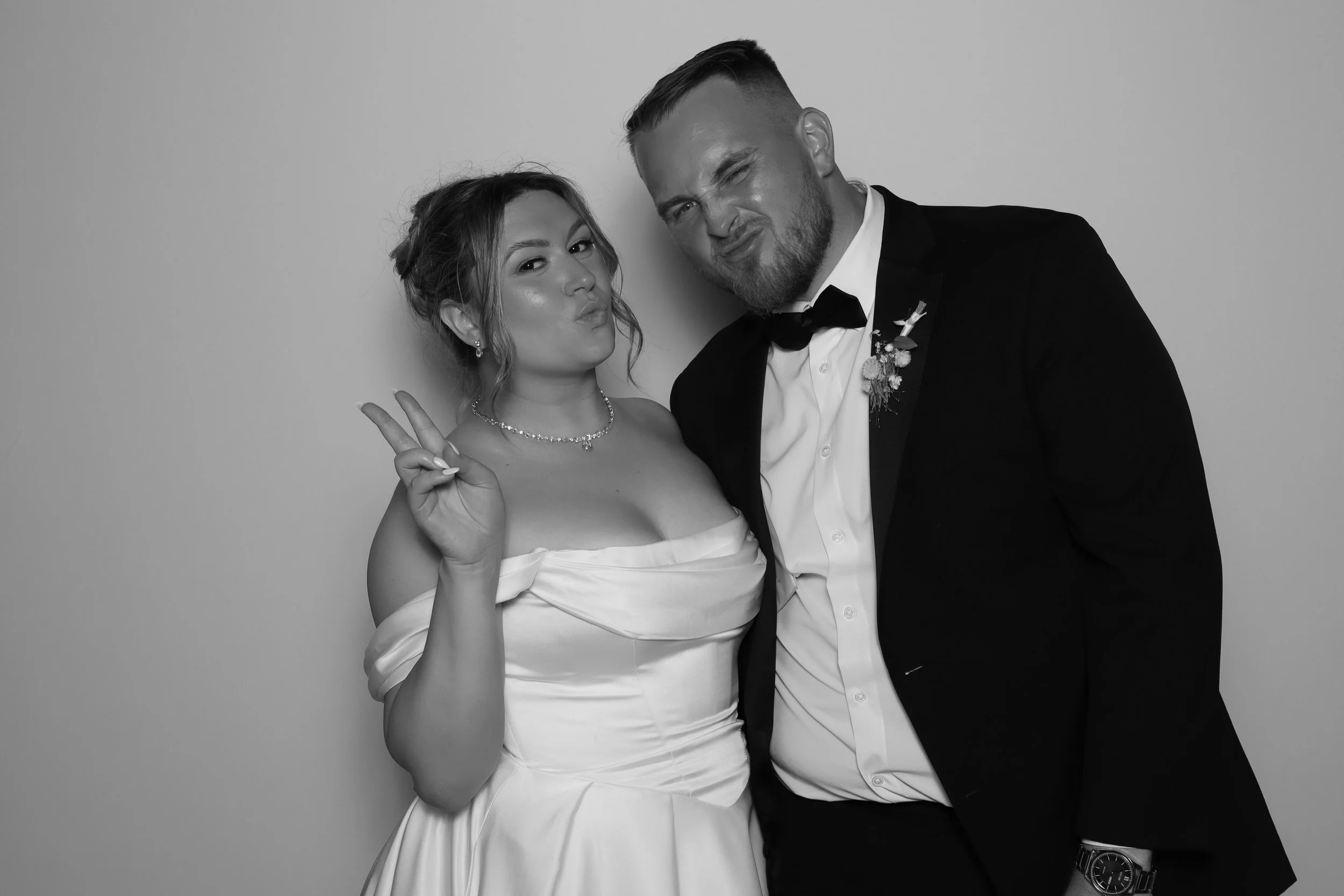 Black and white photo of a bride and groom making silly faces. The bride, wearing a strapless wedding dress with a pearl necklace, holds up a peace sign. The groom, wearing a tuxedo with a bow tie and boutonniere, scrunches his face. They are standing close together against a plain background.