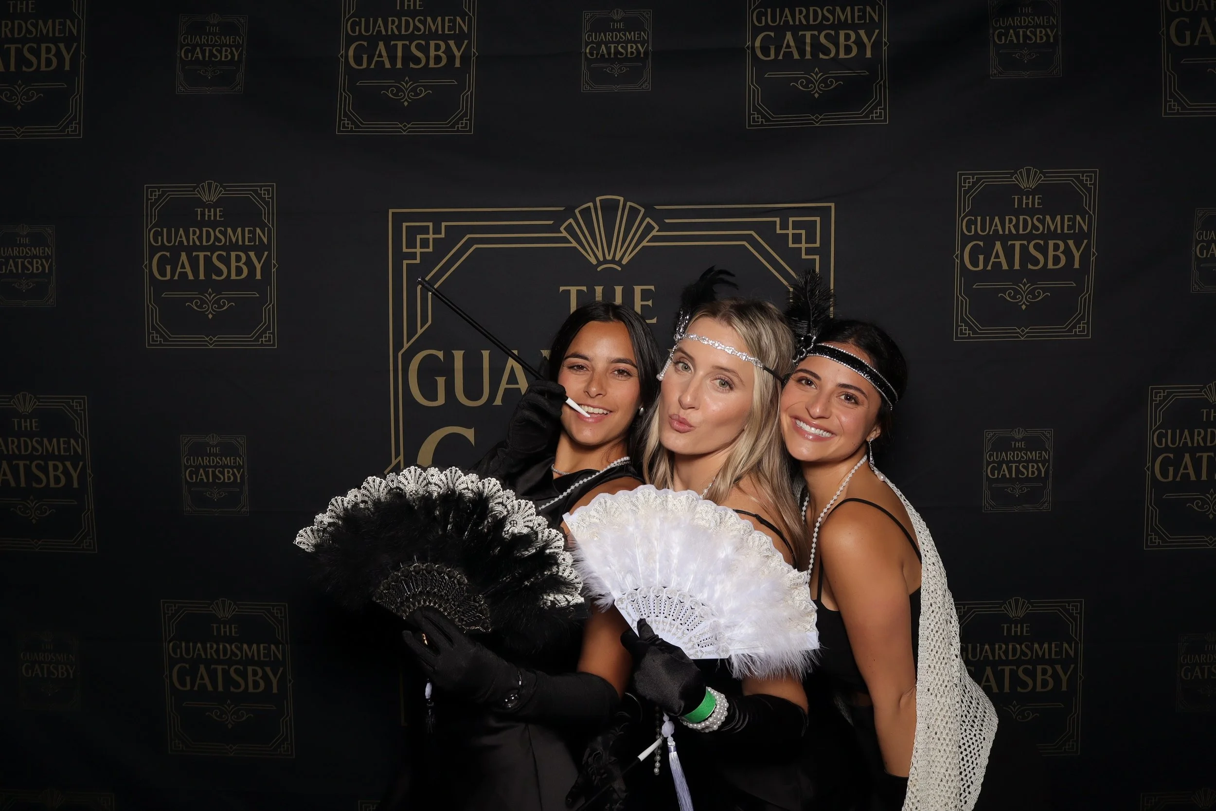 Three women dressed in 1920s flapper costumes, holding fancy fans, standing in front of a backdrop with 'The Guardsmen Gatsby' logo, smiling and posing for the camera.