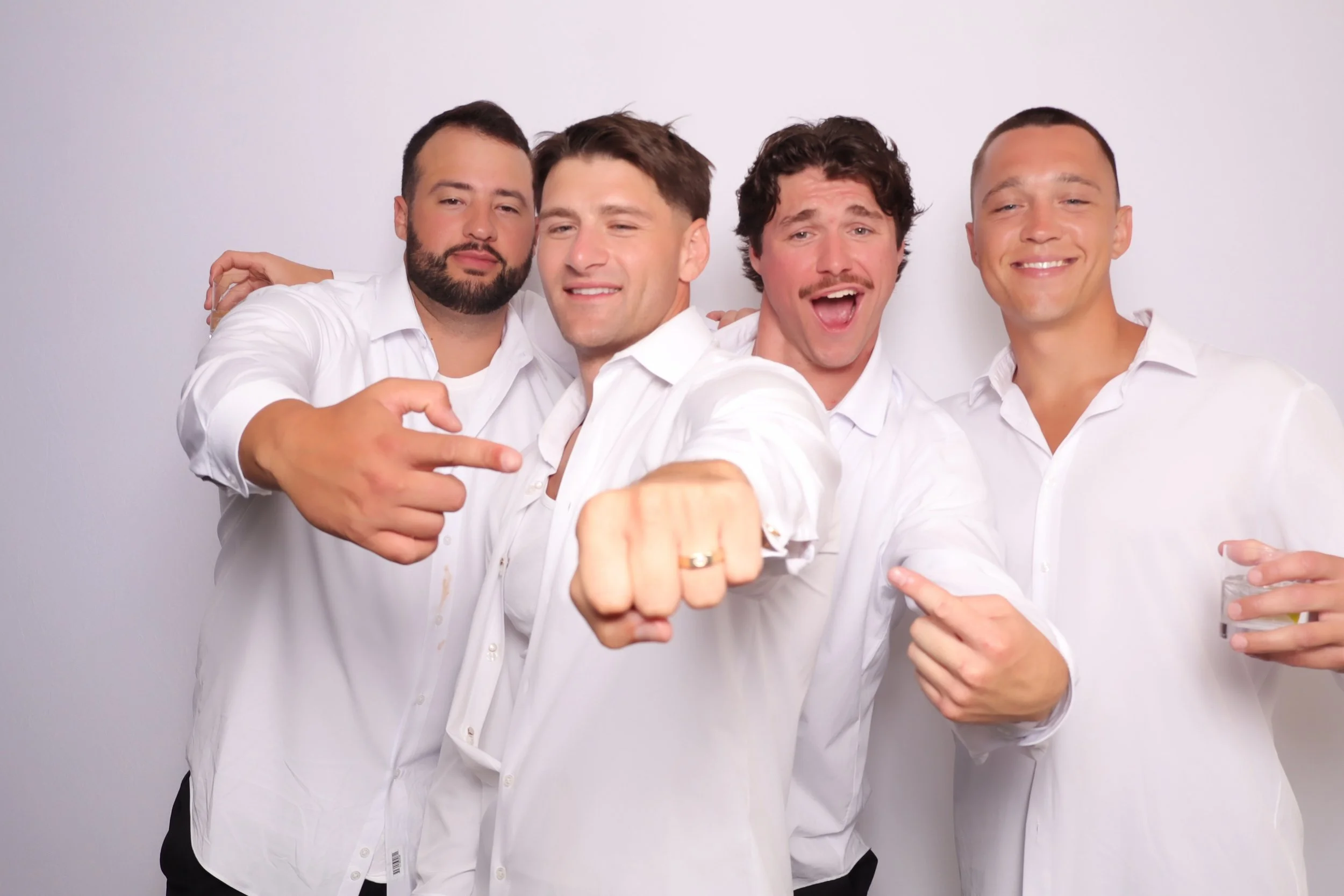Group of four men in Downtown San Diego in white shirts posing together, with one man reaching out to show off his wedding ring, smiling and pointing toward the camera, against a plain white background.