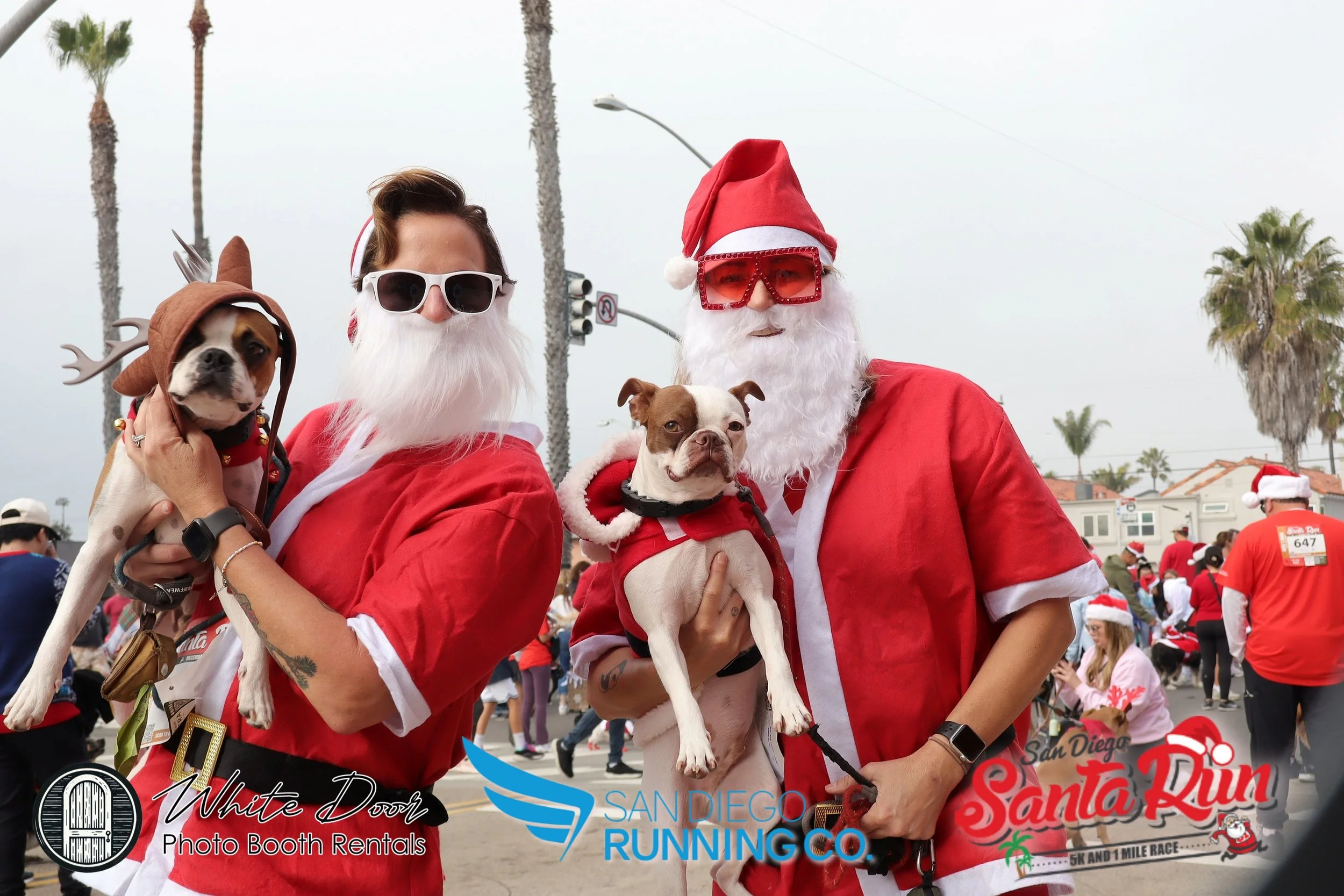 Two people dressed as Santa Claus, each holding a dog, at a holiday race event, with other participants in the background.