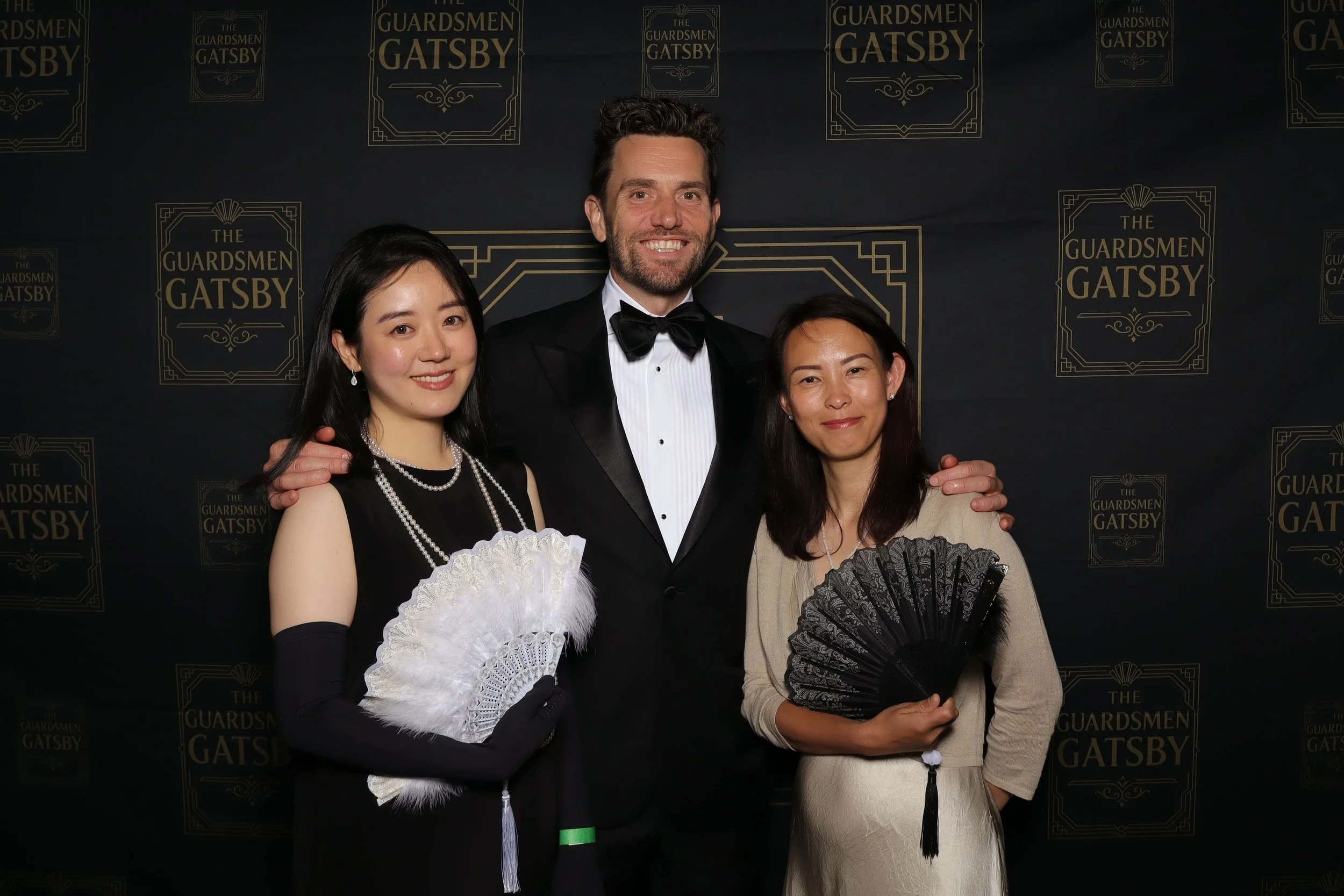 Three people in formal attire posing at an event with a backdrop featuring the title "The Guardsmen Gatsby" in gold lettering on black.