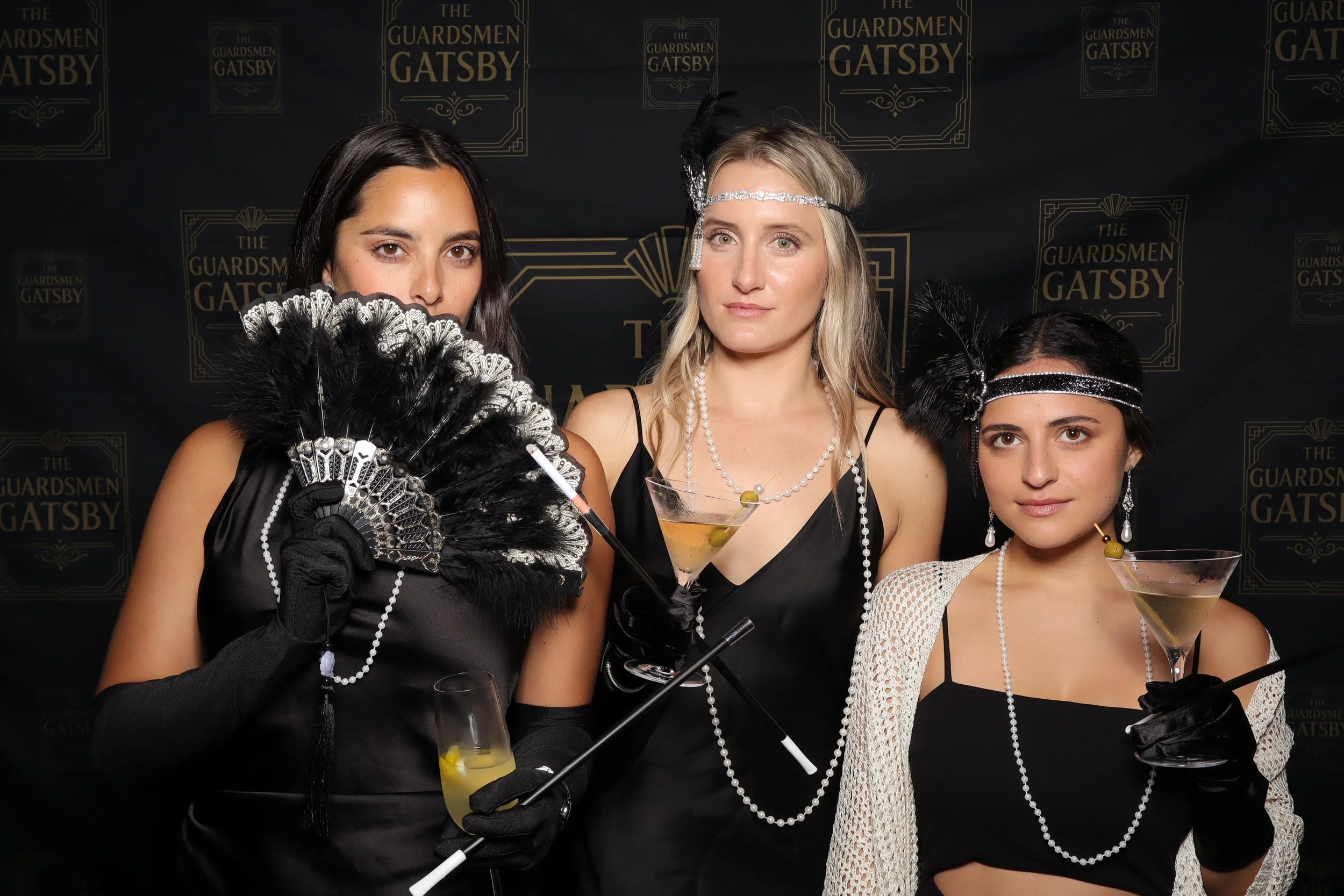 Three women dressed in 1920s flapper costumes pose at a themed party, with a backdrop reading 'The Guardsman Gatsby', holding cocktail drinks, wearing headbands, pearls, gloves, and feather accessories.