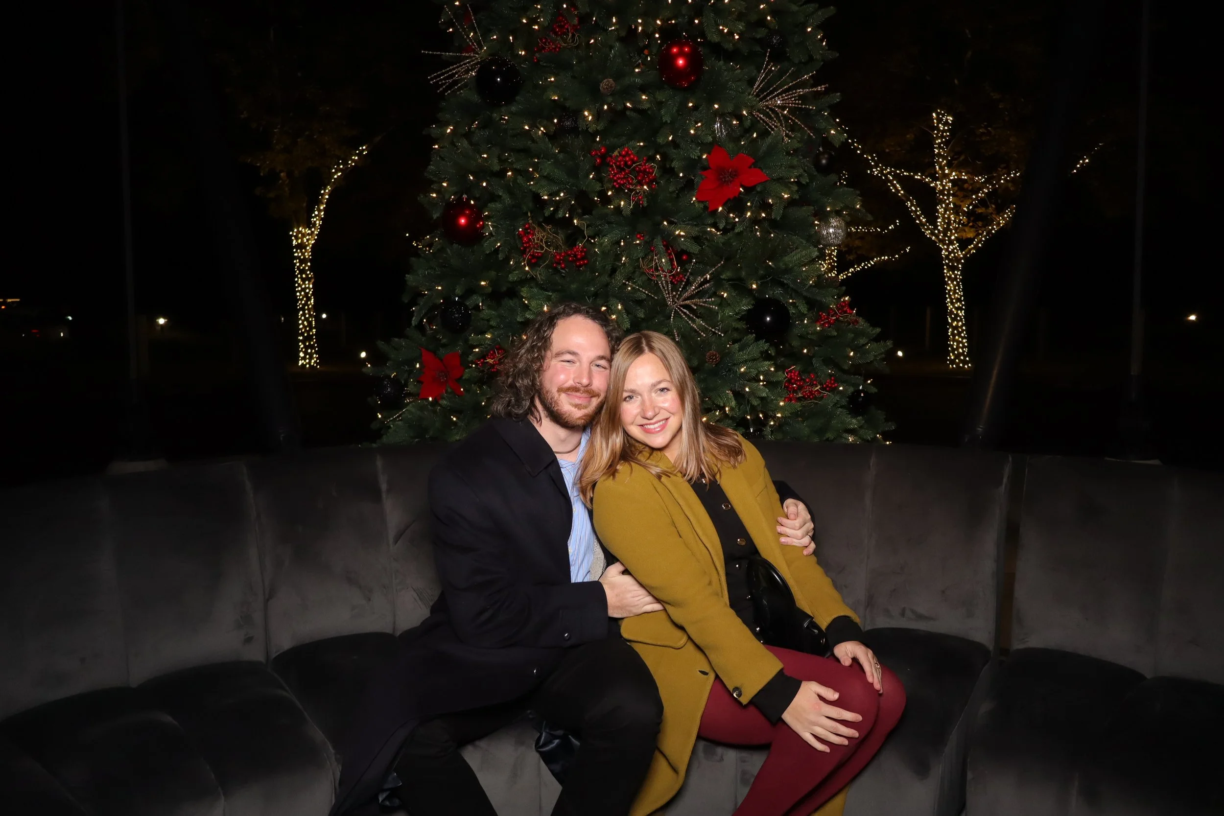 Two people sitting on a dark gray velvet couch in front of a decorated Christmas tree at night. The man has shoulder-length curly hair, a beard, and is wearing a black jacket over a light blue shirt. The woman has shoulder-length blonde hair, is smiling, and is wearing a mustard yellow coat and maroon pants. The Christmas tree behind them is decorated with red poinsettias, berries, black and silver ornaments, and string lights.