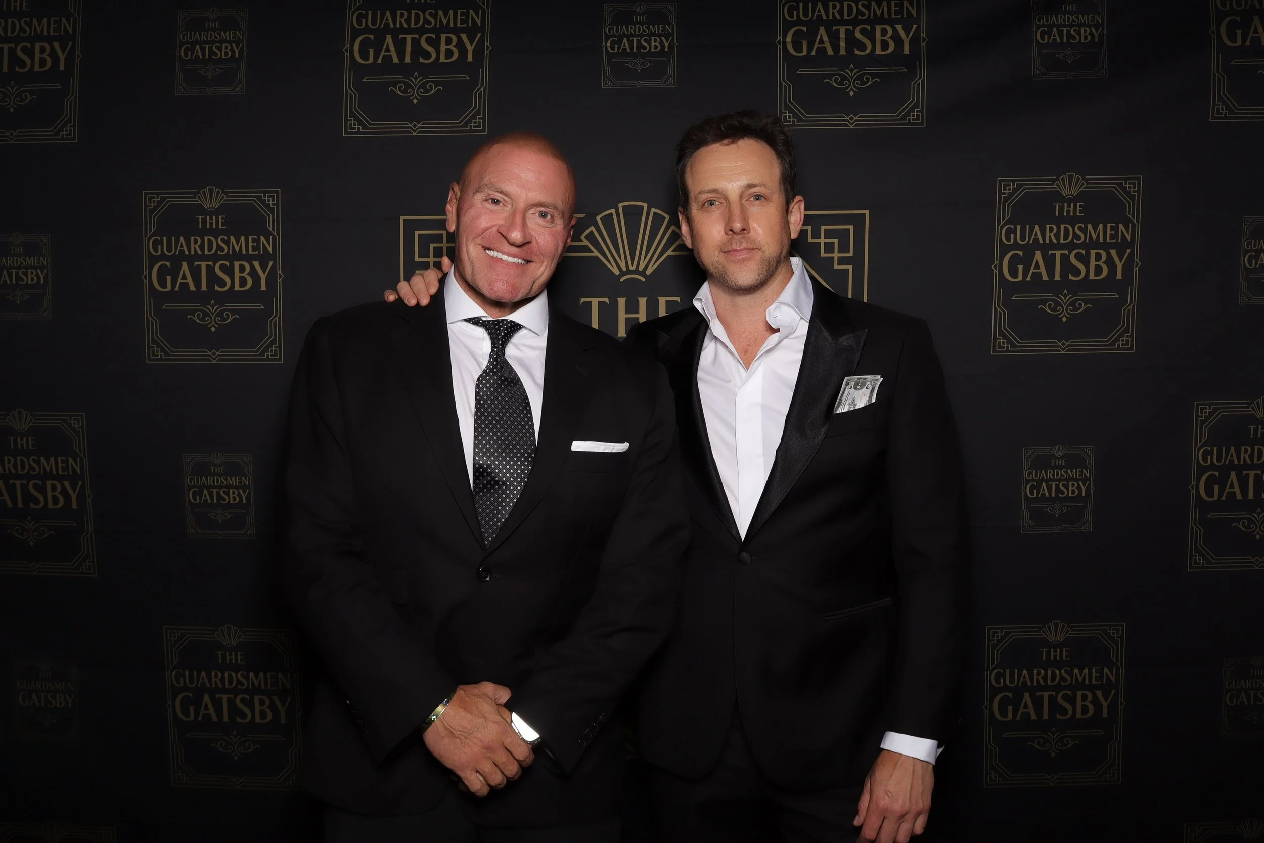 Two men in tuxedos standing side by side in front of a black backdrop with gold lettering that reads "The Guardsmen Gatsby." The man on the left has a shaved head, is smiling, and is holding his hands together in front of him. The man on the right ha