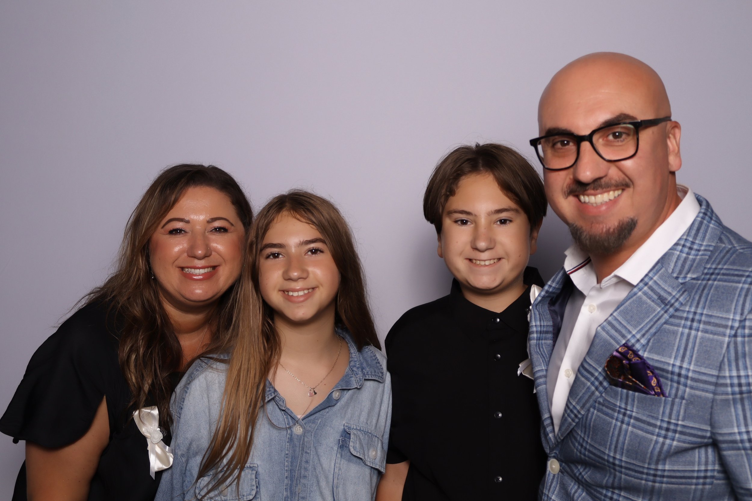 A smiling family of four posing together against a plain background, including a woman, a young girl, a young boy, and a bald man wearing glasses and a blue blazer.