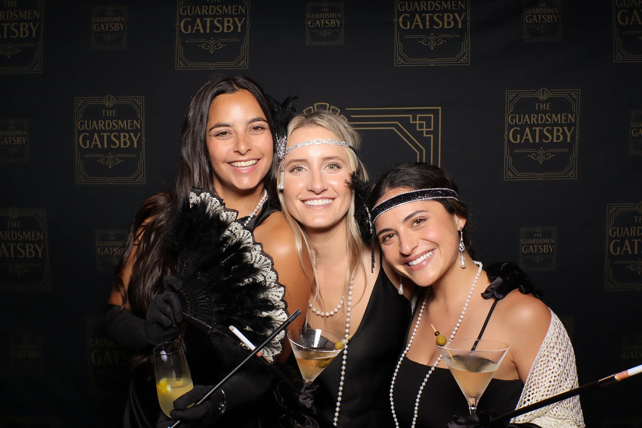 Three women dressed in 1920s style flapper costumes, smiling, holding cocktails, in front of a backdrop with 'The Guardsmen Gatsby' logo.