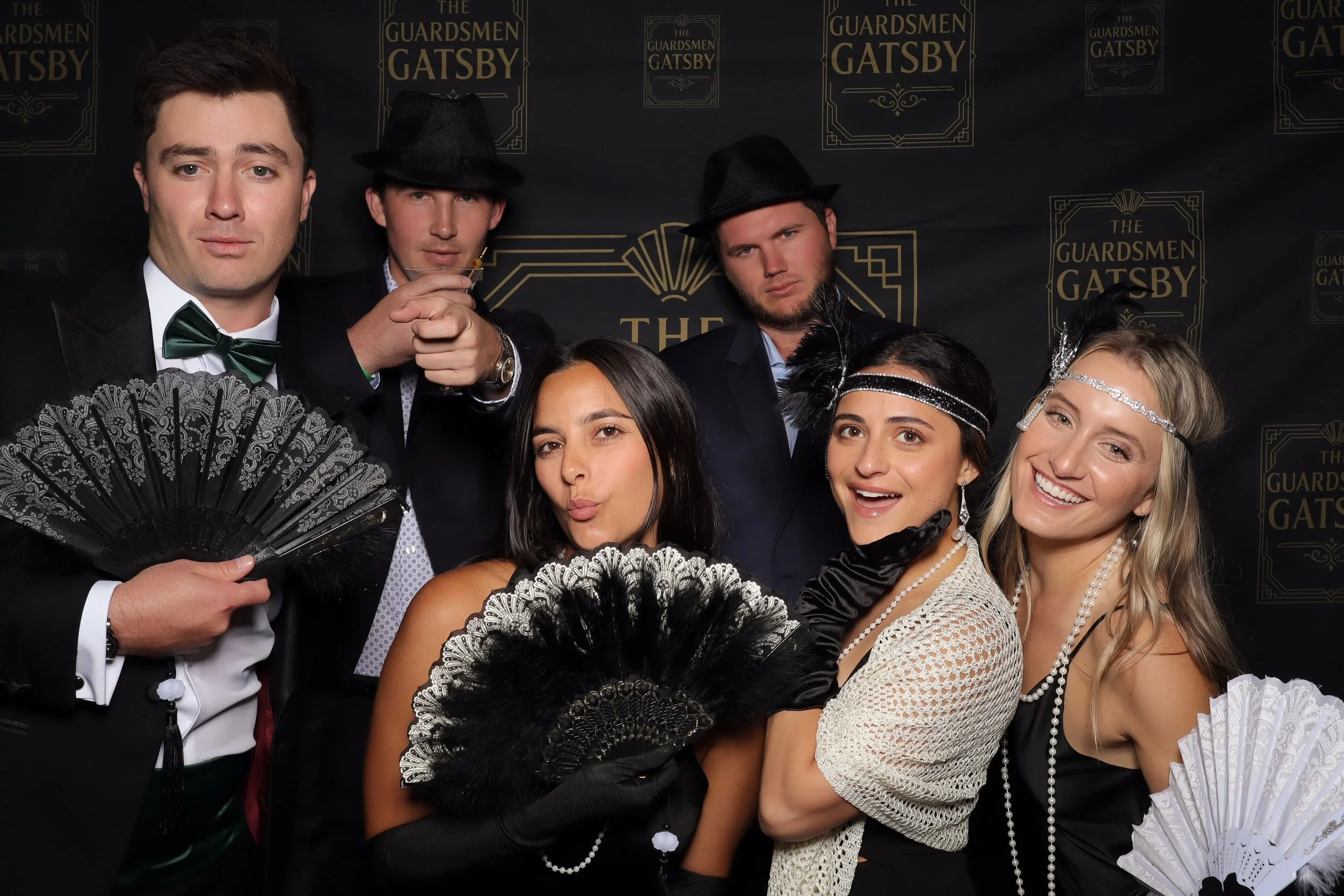 Group of six people dressed in 1920s attire, posing in front of a black backdrop with gold text reading 'The Guardsmen Gatsby'. Three men are wearing suits, hats, and bow ties, and three women are wearing vintage dress, headbands, and gloves, some ho