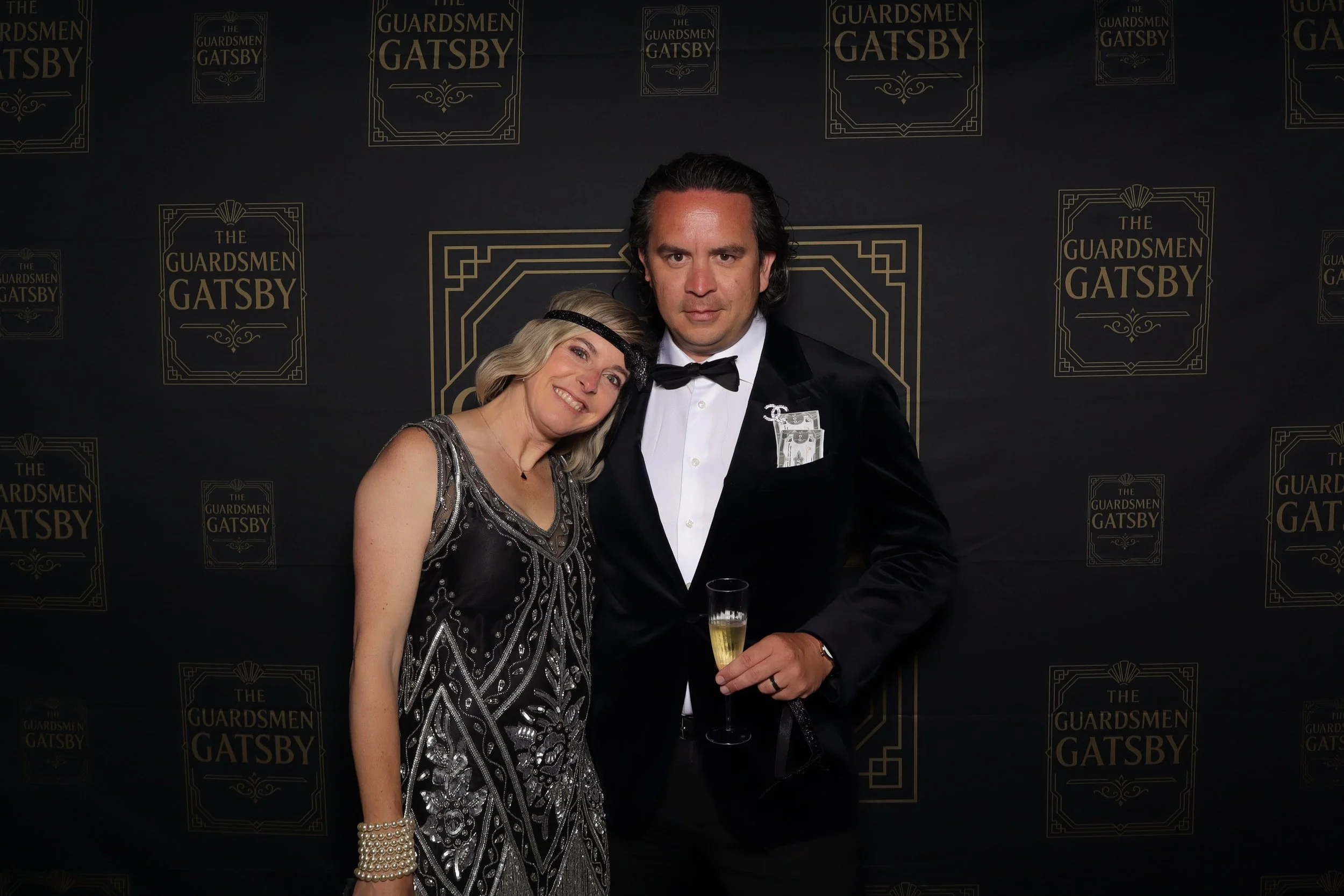 A man and woman dressed in formal attire at a themed event, standing in front of a backdrop with the text 'The Guardsmen Gatsby' and holding a champagne glass.
