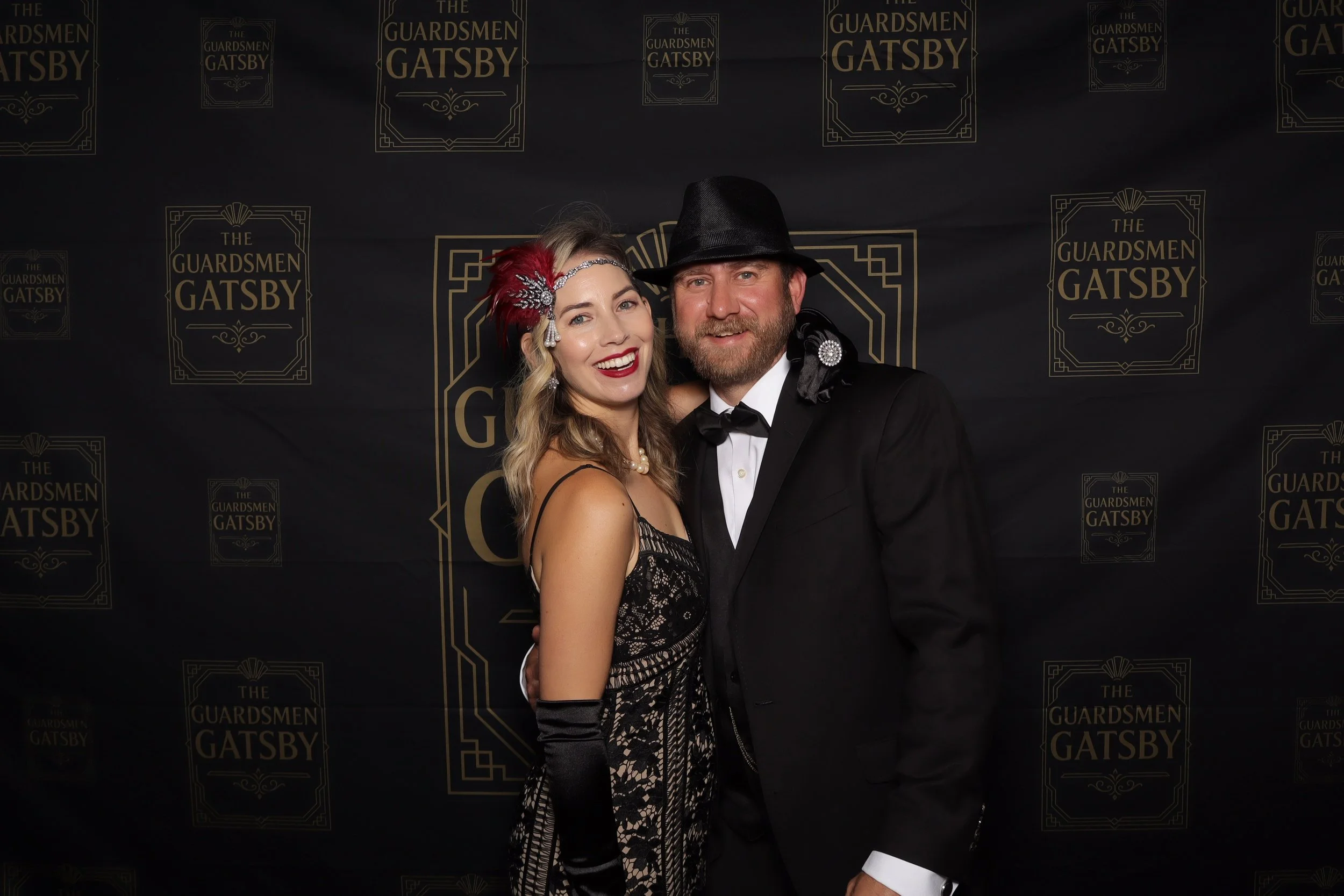 A man in a tuxedo and fedora hat standing next to a woman in a vintage dress with a red feather and hair accessory, both smiling at a black and gold 'The Guardsmen Gatsby' themed event backdrop.