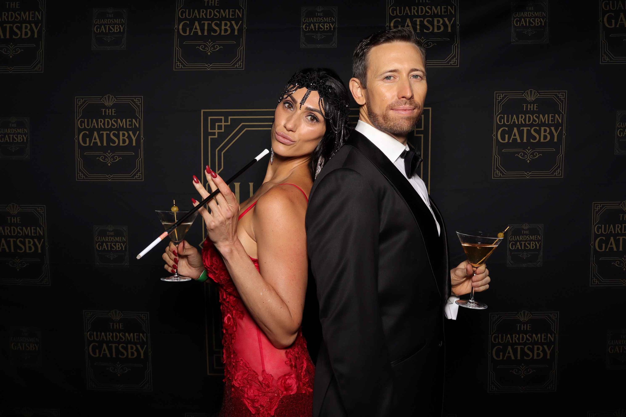 Two people standing back-to-back at a black-themed event background. The woman on the left is wearing a red dress, holding a martini glass and a cigarette holder. The man on the right is wearing a black tuxedo, holding a martini glass. The background features the text "The Guardsmen Gatsby" repeatedly in gold.