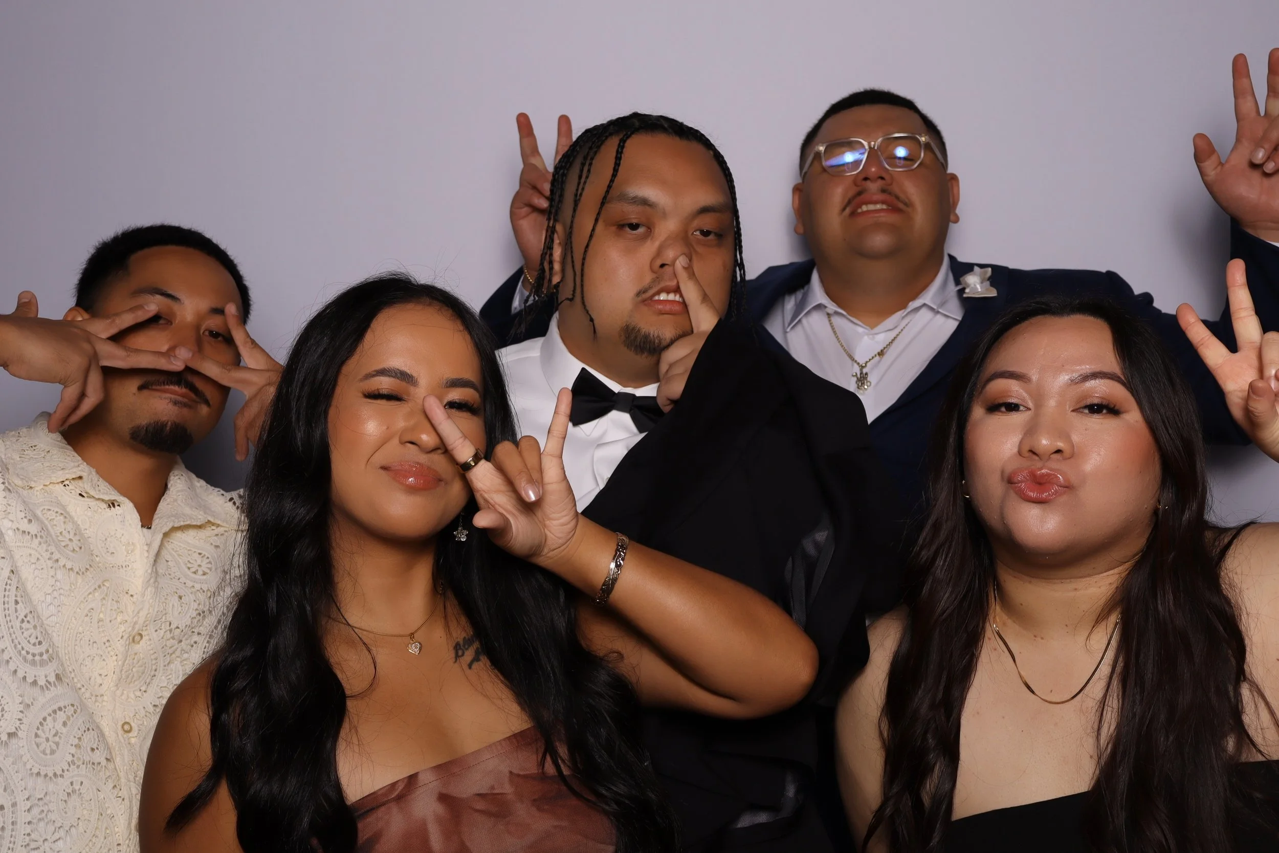 Group of six young adults posing playfully for a photo, some making peace signs or funny faces, against a plain gray background.