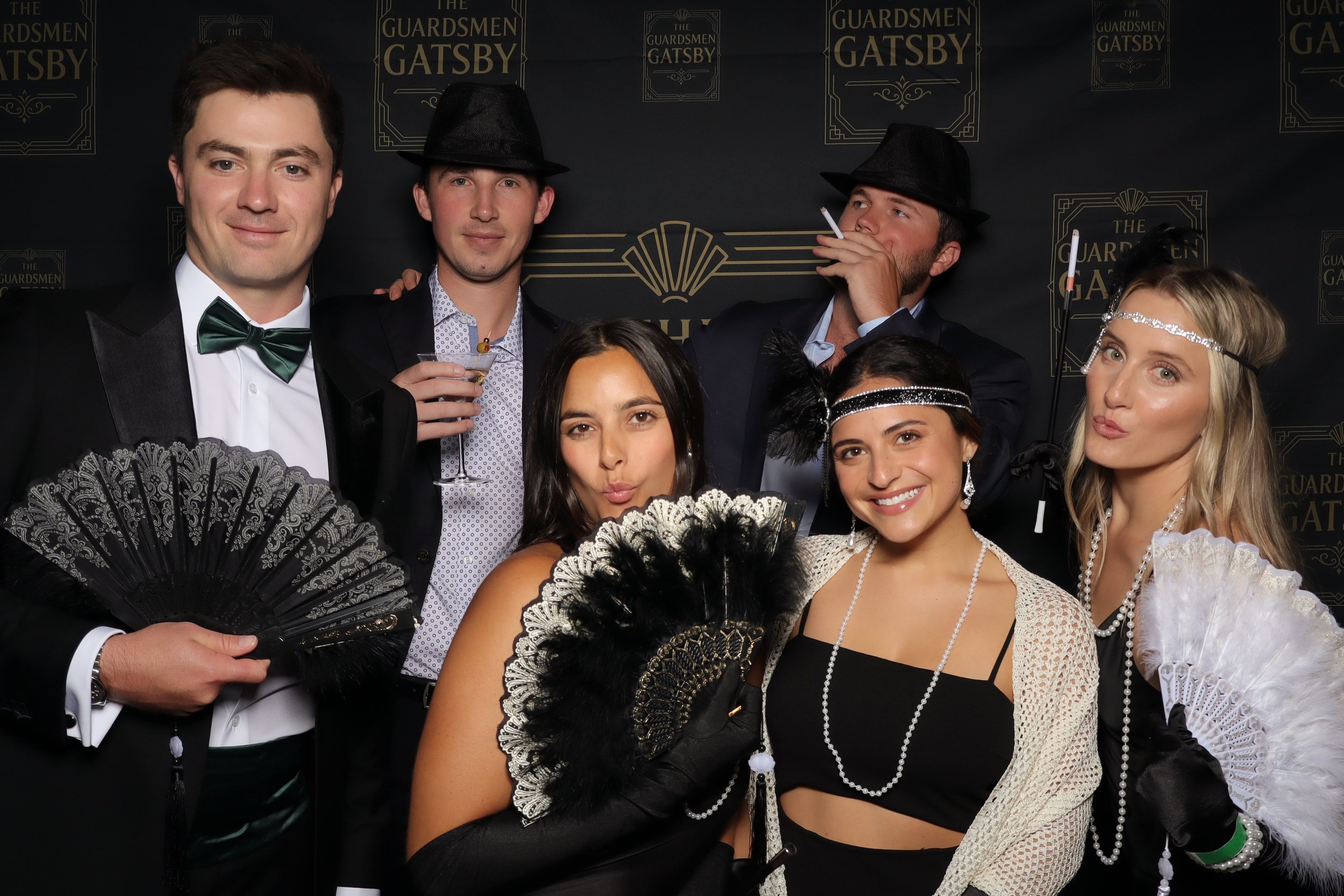 Group of six people at a Gatsby-themed costume party, dressed in 1920s style outfits with accessories such as fans, headbands, and suits, posing in front of a black backdrop with gold writing.