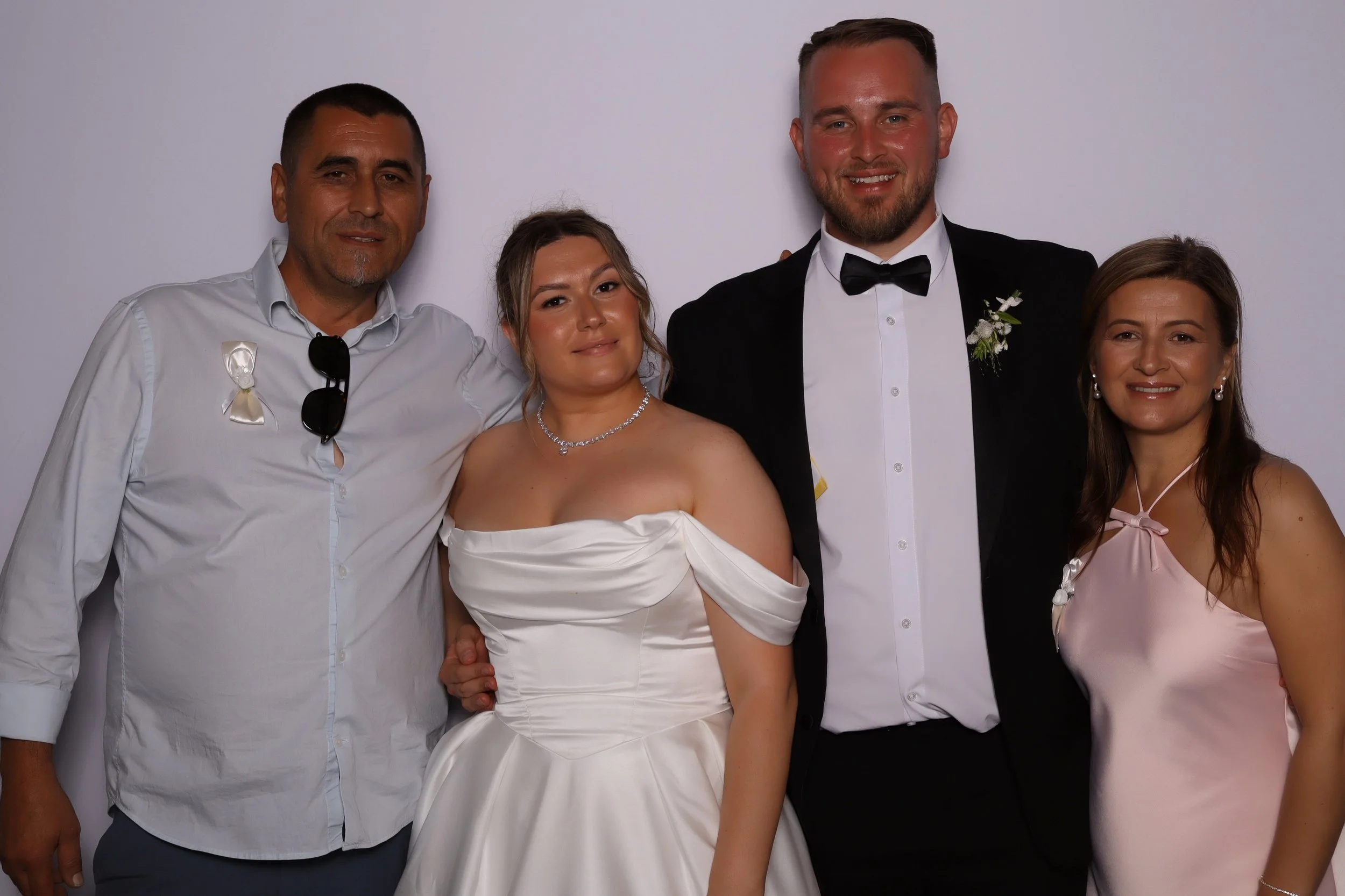 A group of four people dressed in formal attire at a wedding, standing against a plain white background. The man on the left is wearing a light gray shirt with sunglasses hanging from the collar, and a small white ribbon pin. The woman next to him is