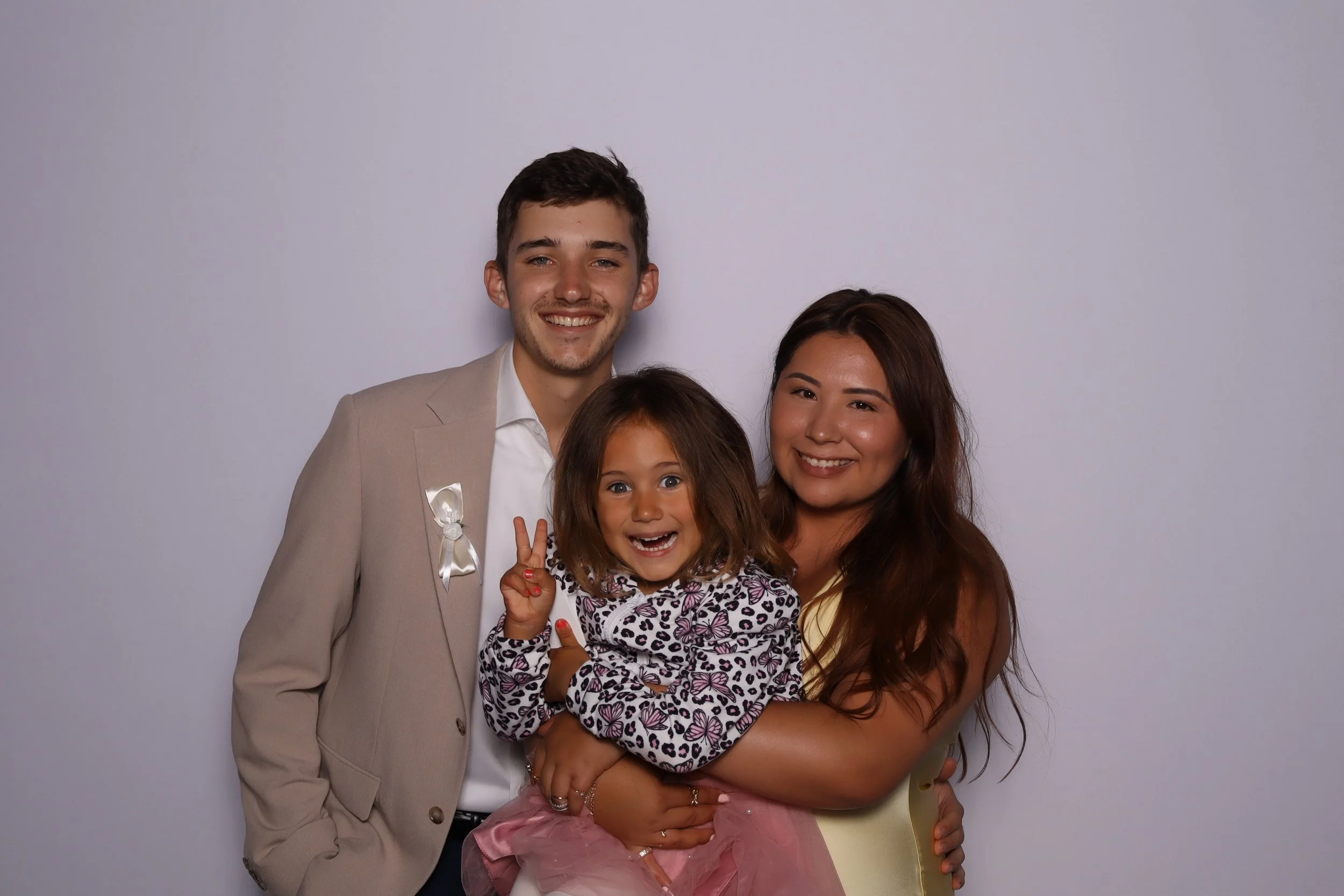 A smiling family of three posing for a photo against a plain, light-colored background. The man is on the left, wearing a beige blazer and white shirt, with his arm around the woman on the right, who has long brown hair and is wearing a light-colored