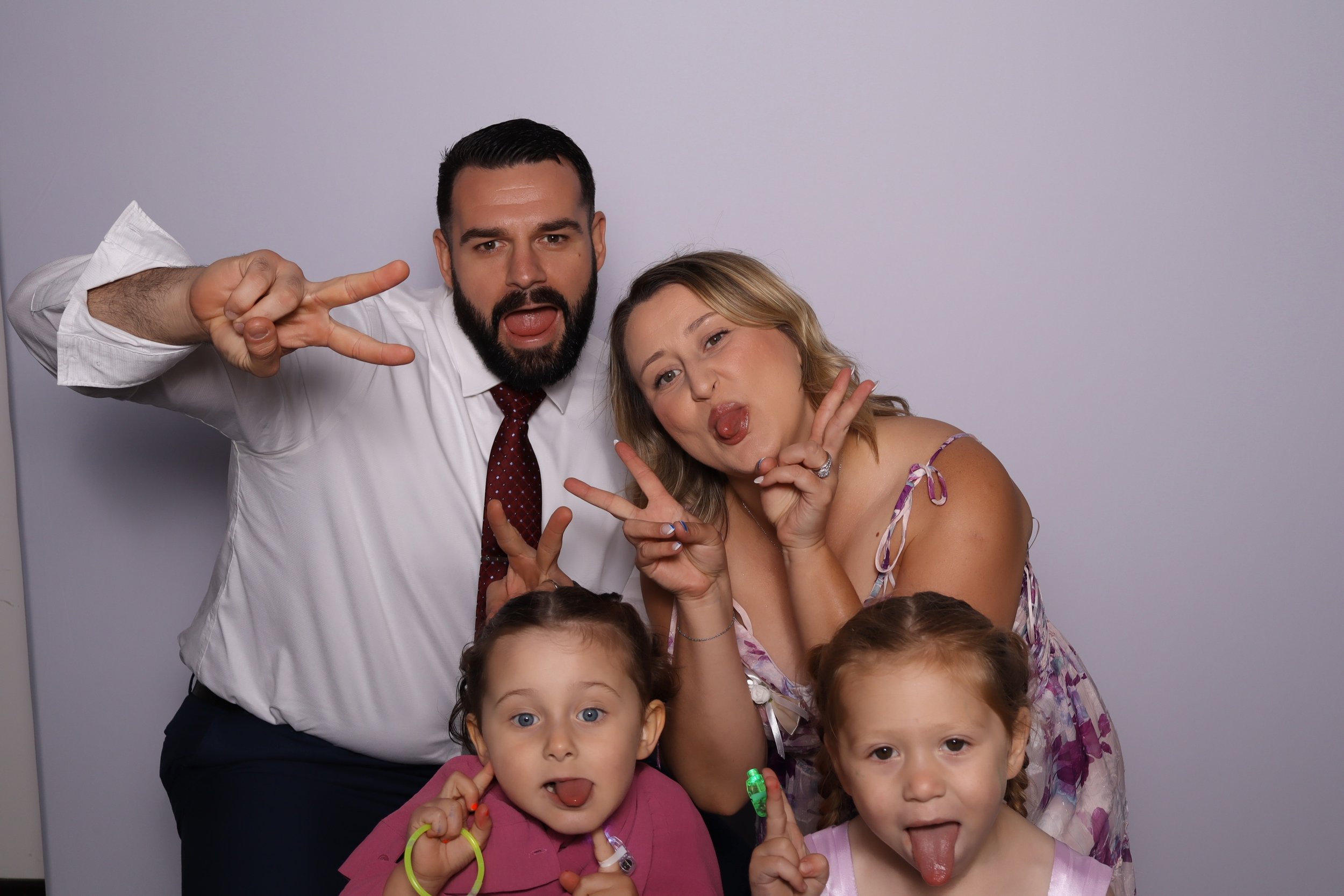 A group of four people, two adults and two children, posing playfully in front of a plain background, sticking out their tongues and making peace signs.