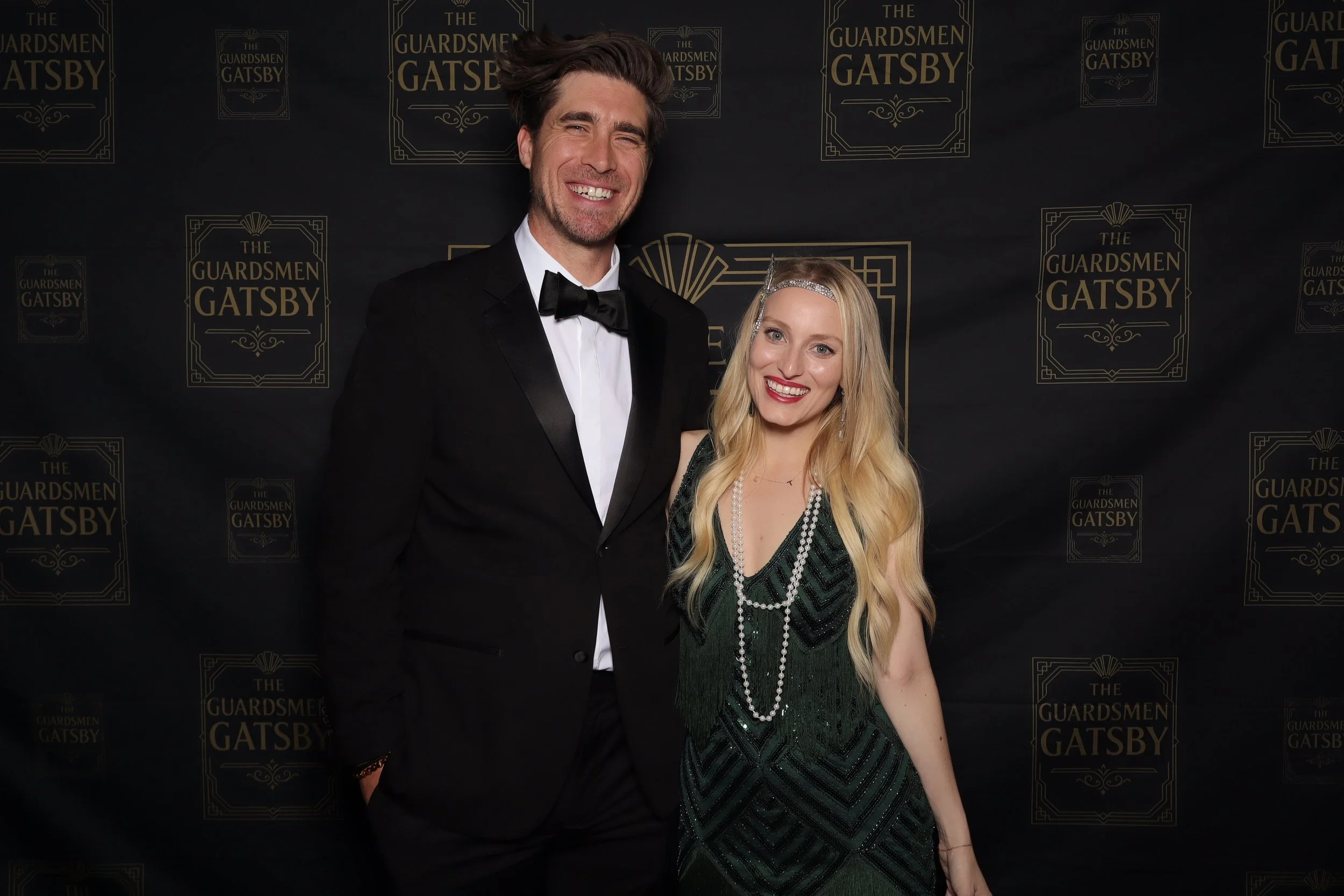 A man and woman dressed in formal attire posing on a black backdrop with gold text and designs for The Guardsmen Gatsby event.