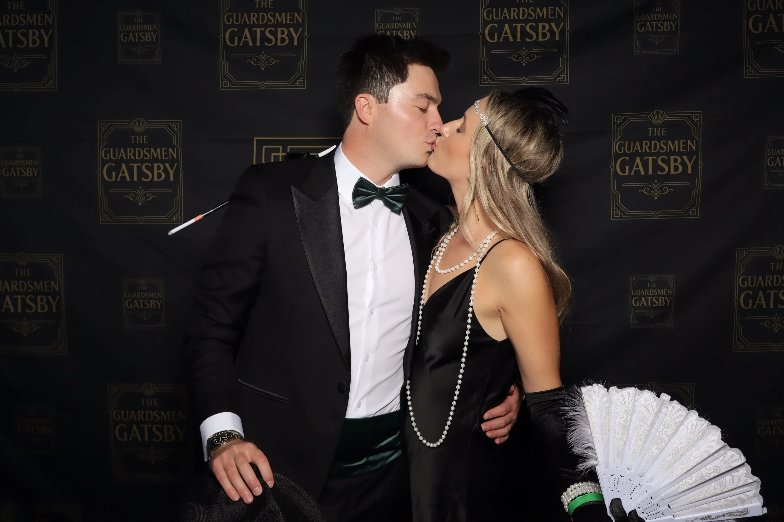 A man and woman dressed in vintage-style attire, kissing in front of a backdrop with 'The Guardsmen Gatsby' text, the man wearing a tuxedo with a bow tie and the woman wearing a black dress with pearl necklaces and a headband, holding a white fan wit