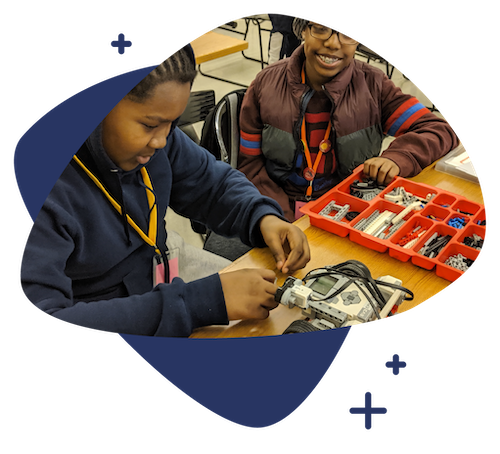 Two young students working on a robotics project with a remote-controlled vehicle and a parts organizer on a classroom table.