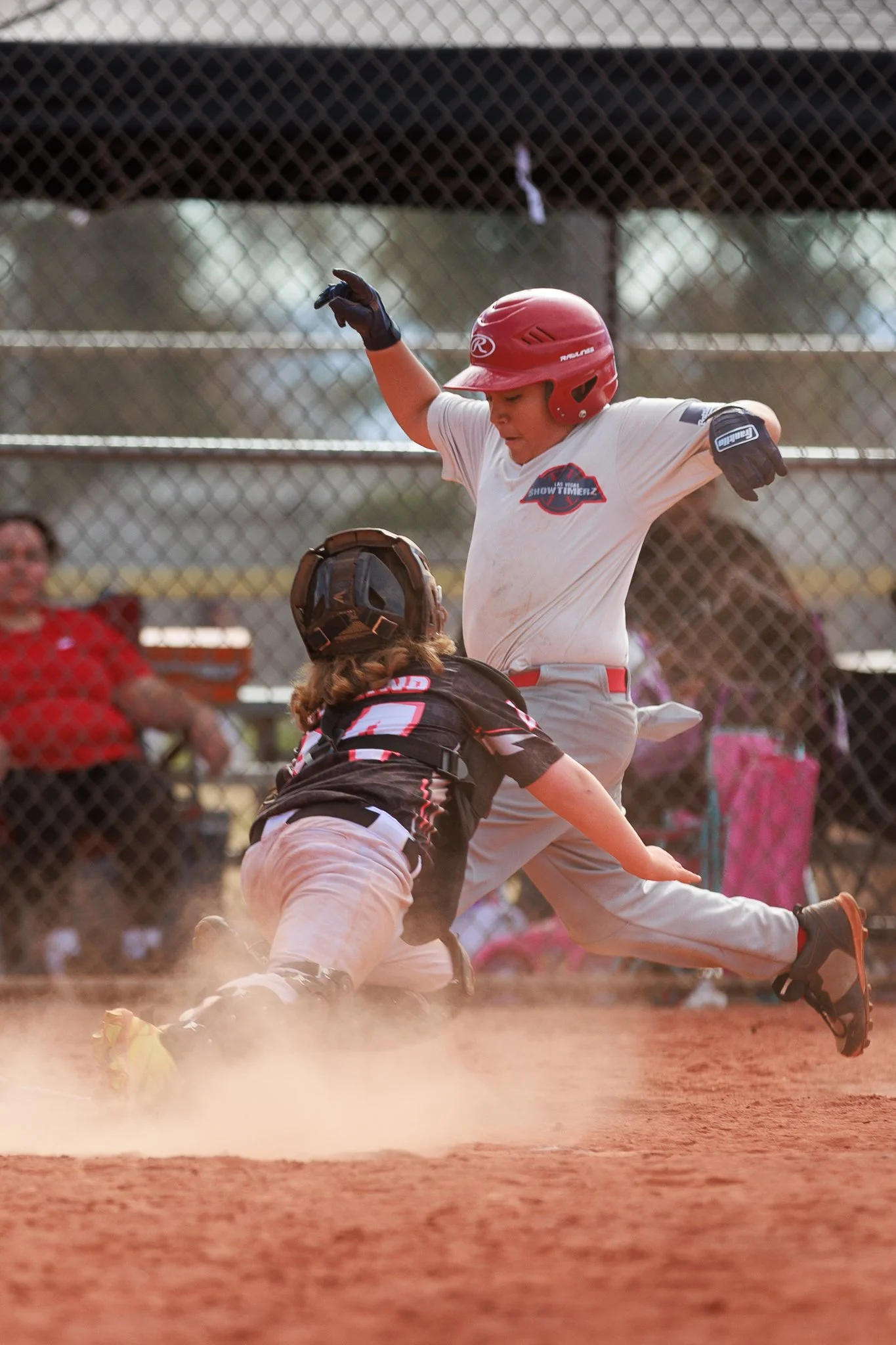 A young baseball player wearing a red helmet and white uniform is sliding into home plate while the catcher, in a black uniform, is attempting to tag him out. Dust is kicked up on the dirt field, and spectators are visible behind a chain-link fence.