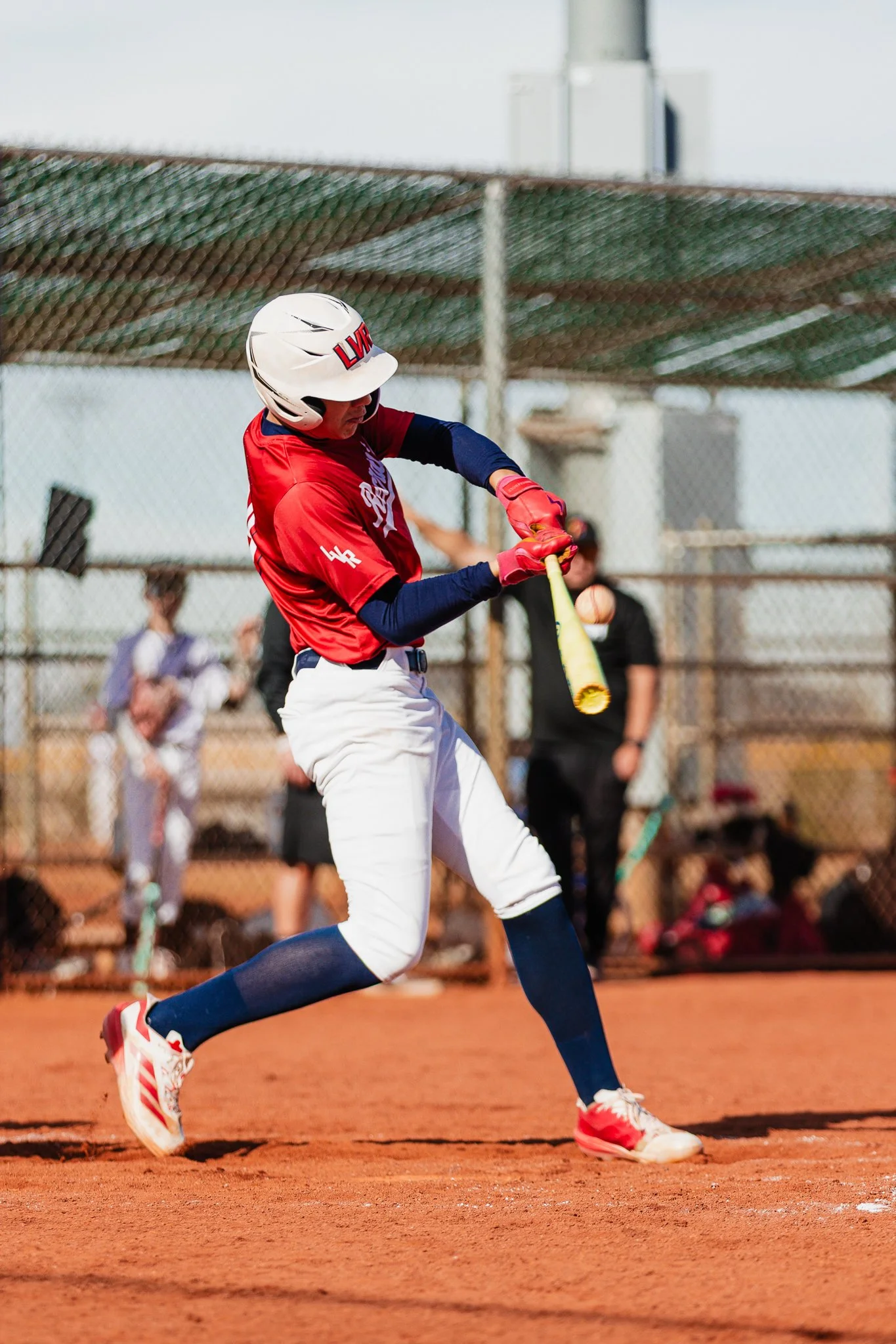 A baseball player in a red jersey and white pants is hitting a baseball on a baseball field. The player is wearing a helmet and red gloves, with a bat in hand, mid-swing. There are spectators and other people in the background.